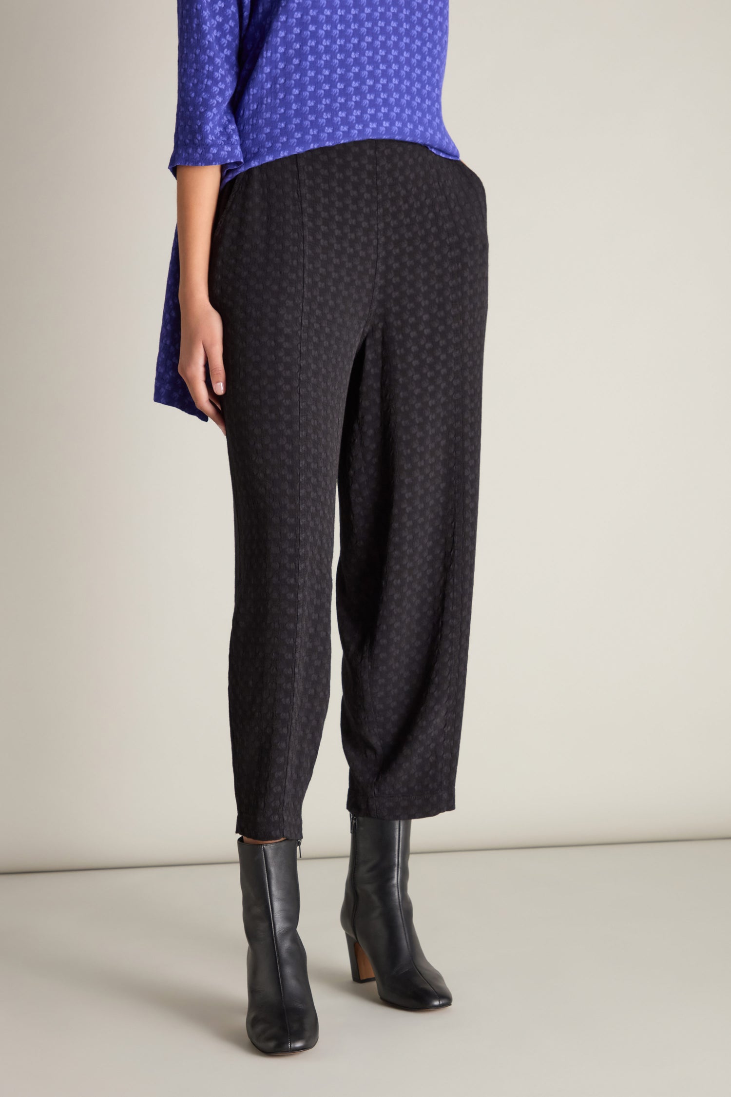 A person is standing against a plain background, wearing a blue patterned top, chic Small Square Jacquard Bubble Trousers with an elasticated waistband, and black ankle boots.