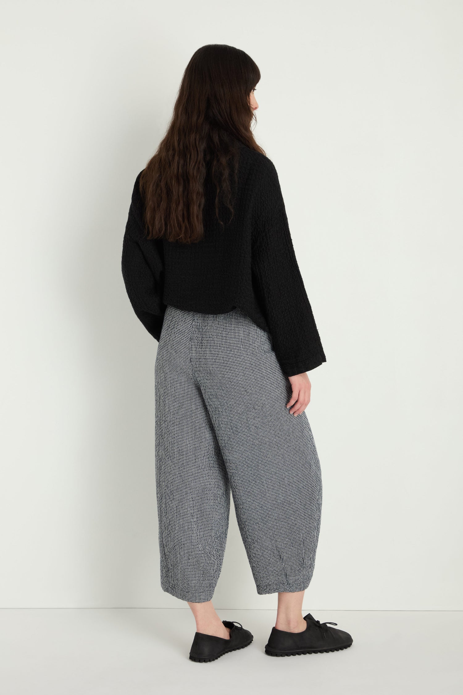 A person with long brown hair faces away, wearing the MTP5167-MGI black textured top, Micro Gingham Bubble Trouser in gray, and black slip-on shoes against a plain white background.