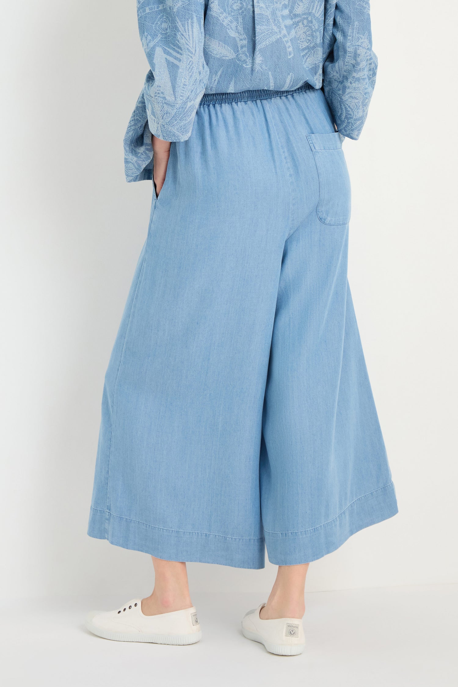 Model wears Fluid Denim Wide Trousers in light blue with a relaxed, wide-leg fit, paired with white slip-on shoes and a blue patterned top against a plain background.