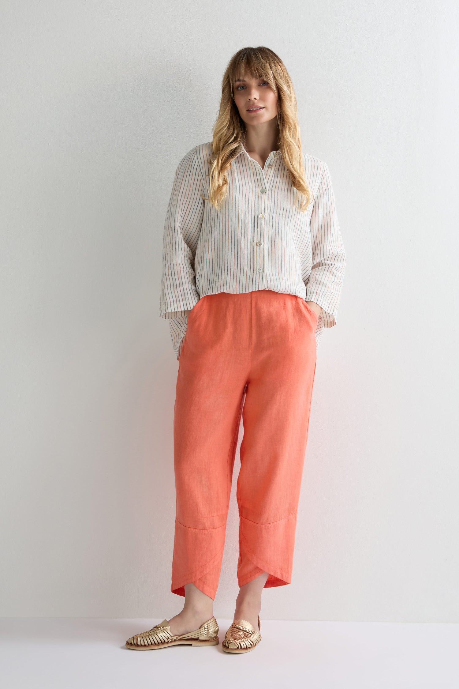 A woman stands against a white wall, wearing a striped button-up shirt and chic coral Linen Tulip Hem Bubble Trousers, paired with gold loafers and her hands casually tucked in her pockets.