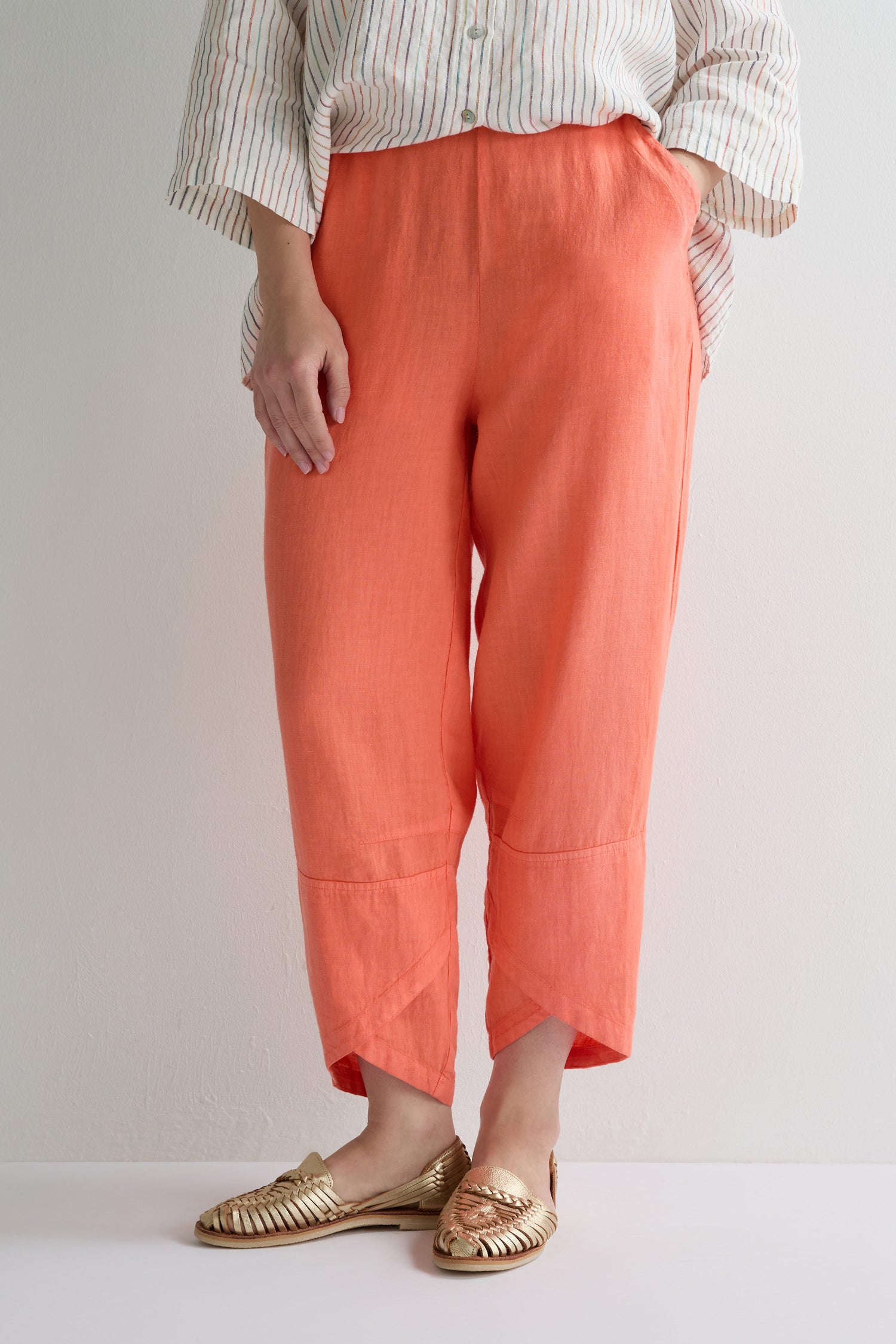 Person wearing coral orange Linen Tulip Hem Bubble Trousers, a striped shirt, and gold slip-on shoes, standing against a plain light-colored background.