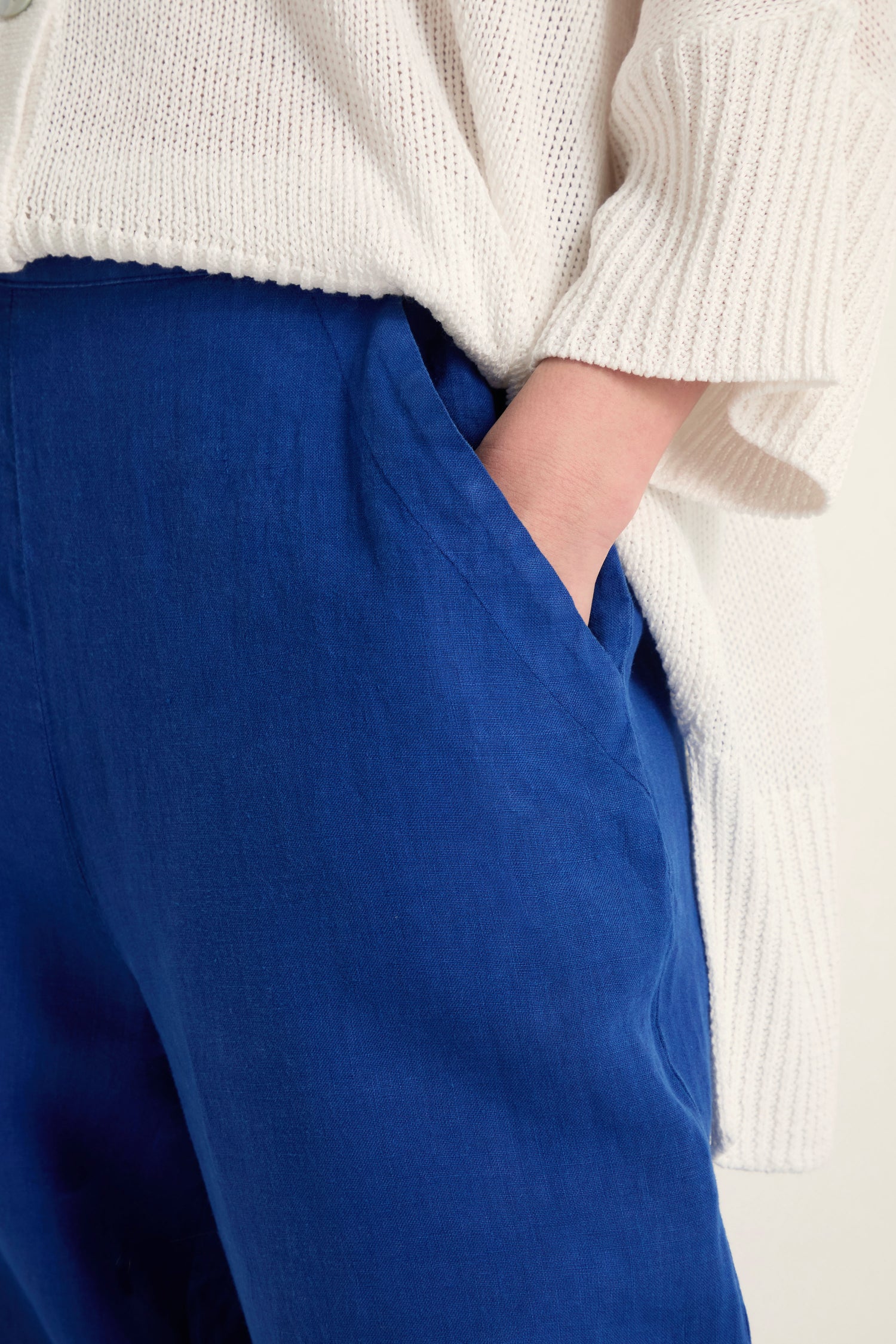 A person wears Linen Tulip Hem Cerulean Bubble Trousers and a cream knit sweater, with one hand in a pants pocket.