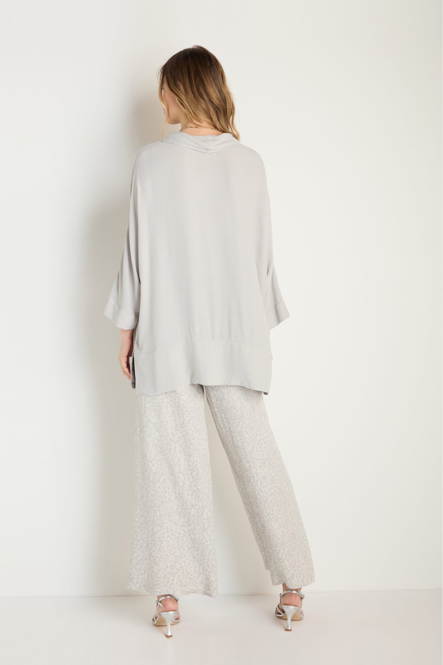 A woman stands facing away from the camera, wearing a loose light gray top, Animal Textured Jacquard Wide Trouser (MTP6715-ATJ), and heeled sandals against a plain white background.