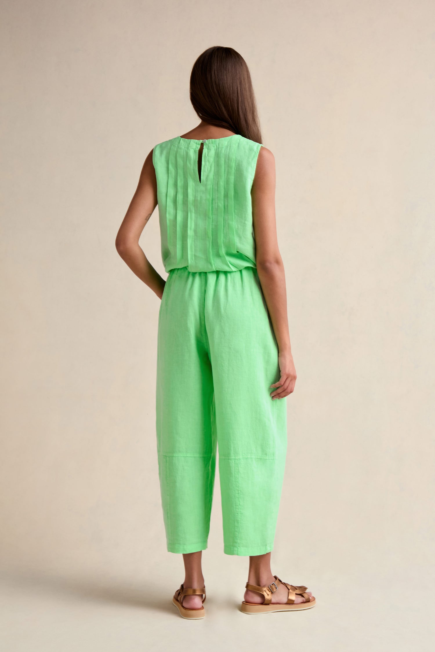 A person stands facing away, wearing Linen Seam Detail Pistachio Bubble Trousers with an elasticated waistband, paired with a sleeveless mint green top and tan sandals, against a plain beige background.