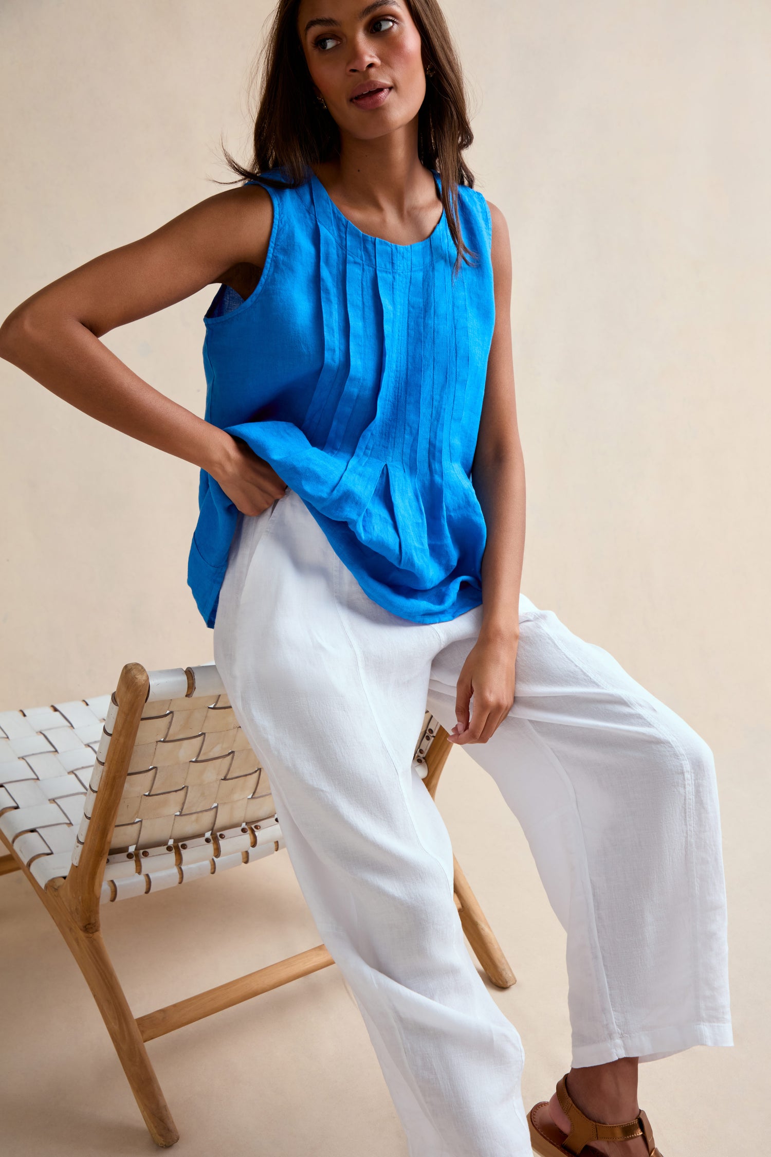 A woman sits on the edge of a woven wooden chair against a beige background, wearing Linen Seam Detail White Bubble Trousers with an elasticated waistband and a bright blue sleeveless pleated top.
