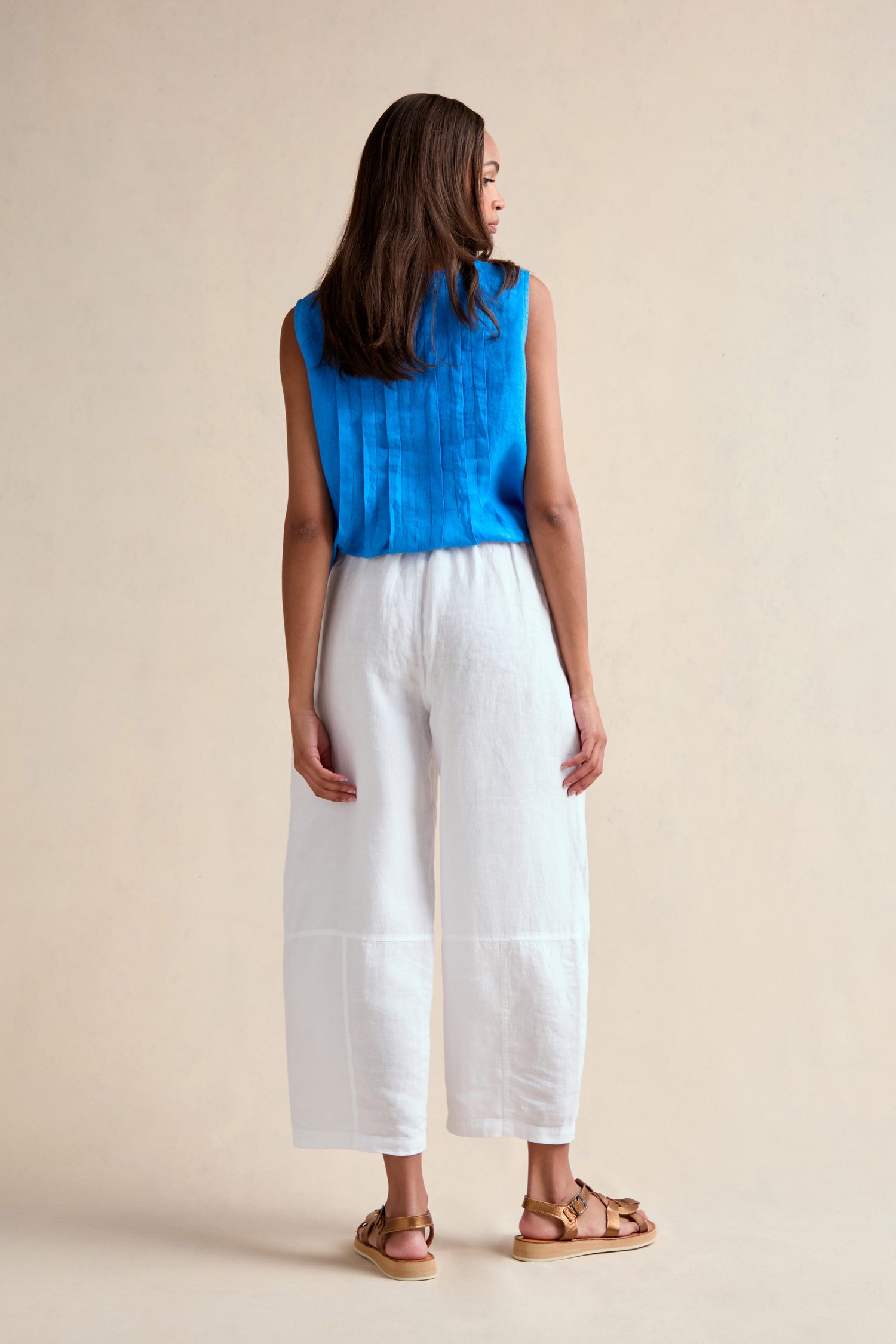 A woman faces away, wearing the Linen Seam Detail White Bubble Trouser with an elasticated waistband and seam details, styled with a sleeveless blue top and tan sandals. The background is plain beige.