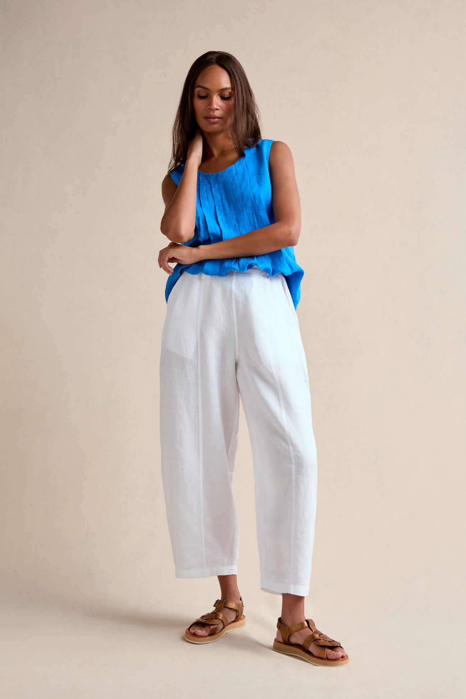 A woman stands against a plain background wearing a blue sleeveless top, the Linen Seam Detail White Bubble Trouser with an elasticated waistband, and brown sandals, one hand touching her neck and the other arm crossed.