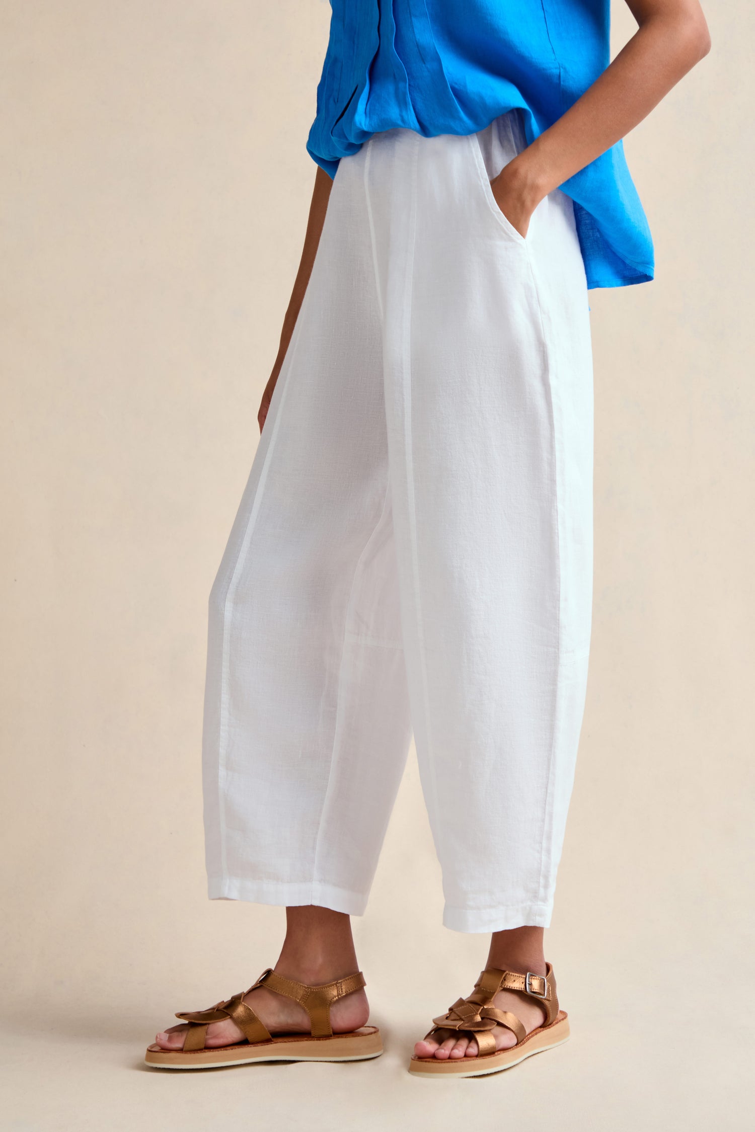 Person wearing a bright blue sleeveless top, Linen Seam Detail White Bubble Trouser with an elasticated waistband, and tan flat sandals, standing with one hand in their pocket.