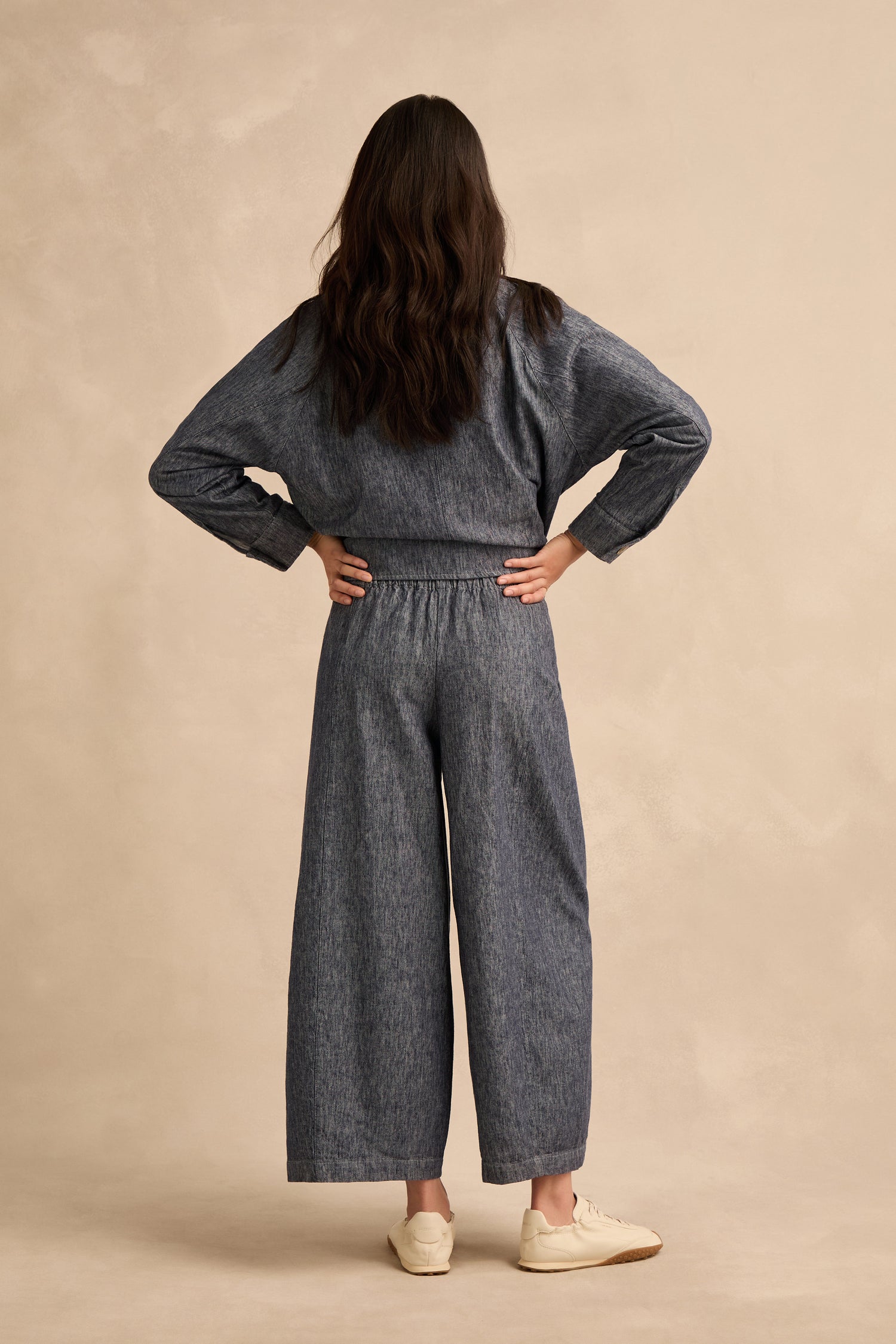 A person stands facing away, hands on hips, wearing a long-sleeve gray top paired with Washed Cotton Linen Barrel Trousers featuring an elasticated waist and white sneakers against a neutral background.