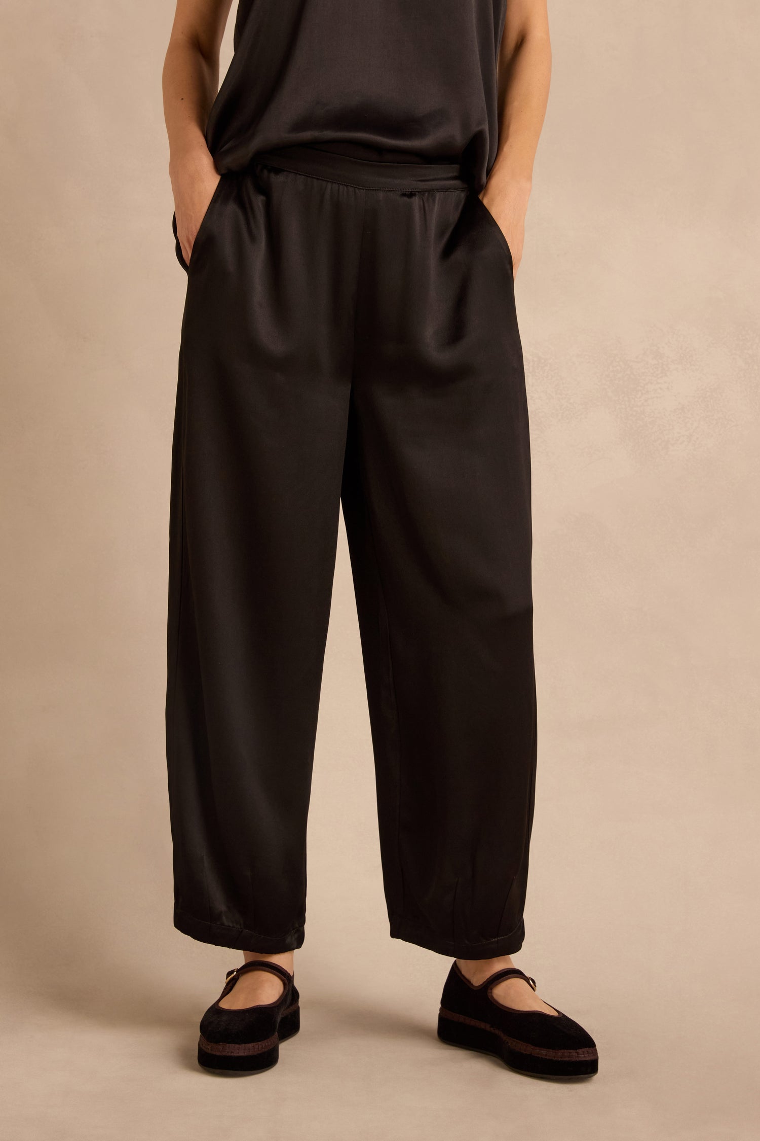 Wearing the Satin Back Crepe Bubble Trousers, a person stands with hands in pockets against a plain beige background, paired with a black sleeveless top and black shoes.