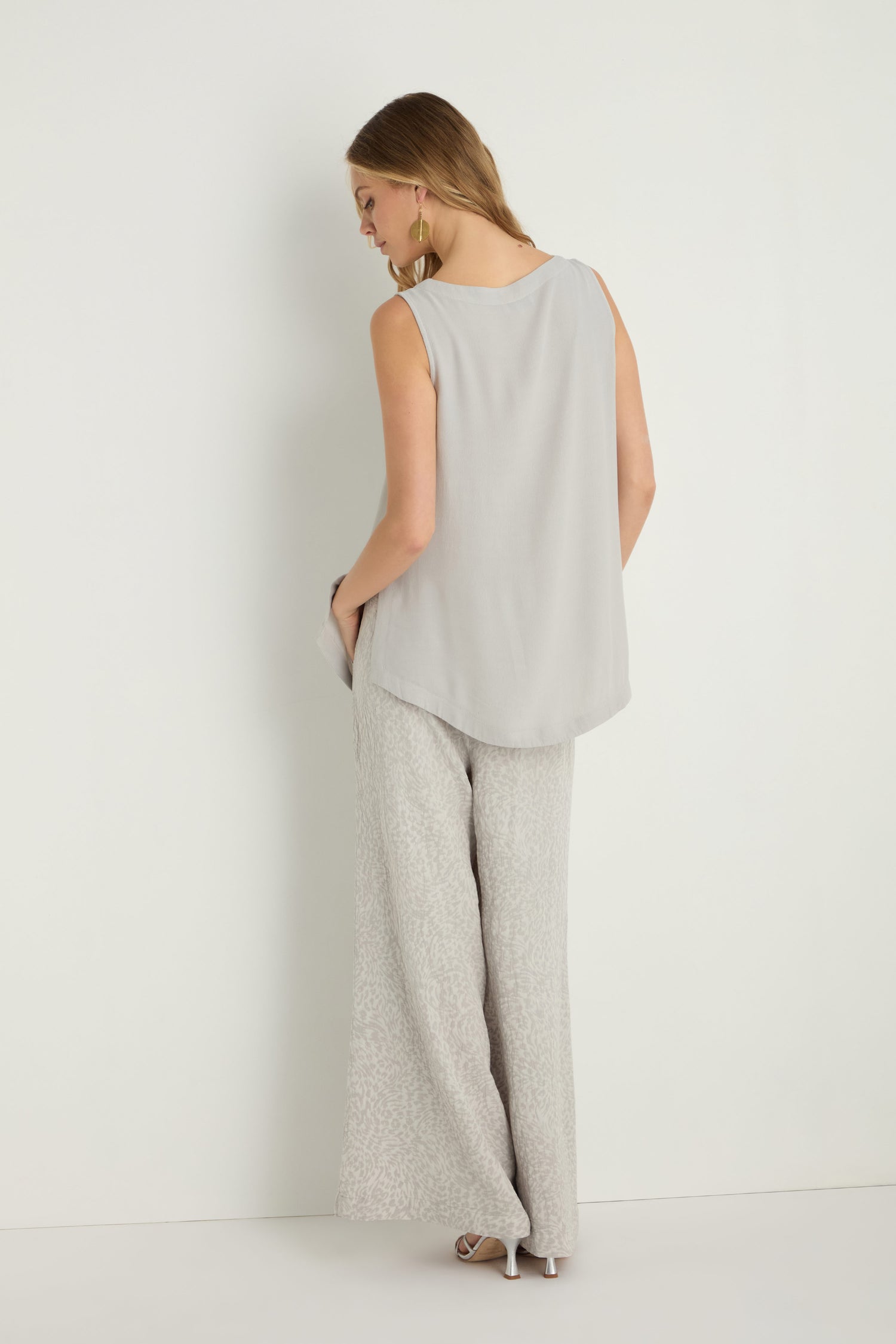 Woman stands against a white wall, facing away, wearing a light gray Crepe Vest—a sleeveless, lightweight layer—paired with wide-leg patterned pants and high-heeled sandals.