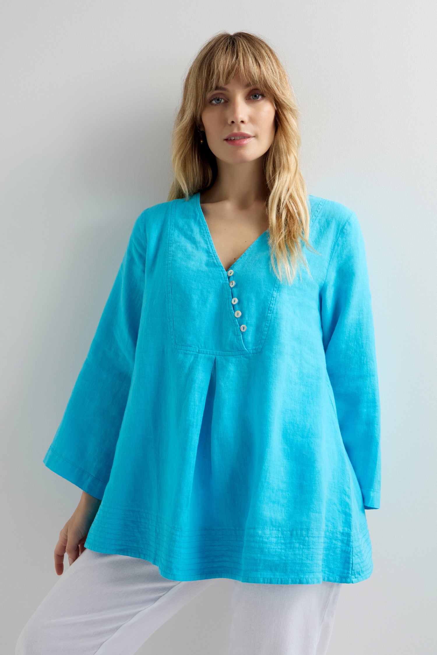 A person stands against a white background in a vibrant blue Linen Button Placket Tunic with white pants, highlighting its relaxed fit.