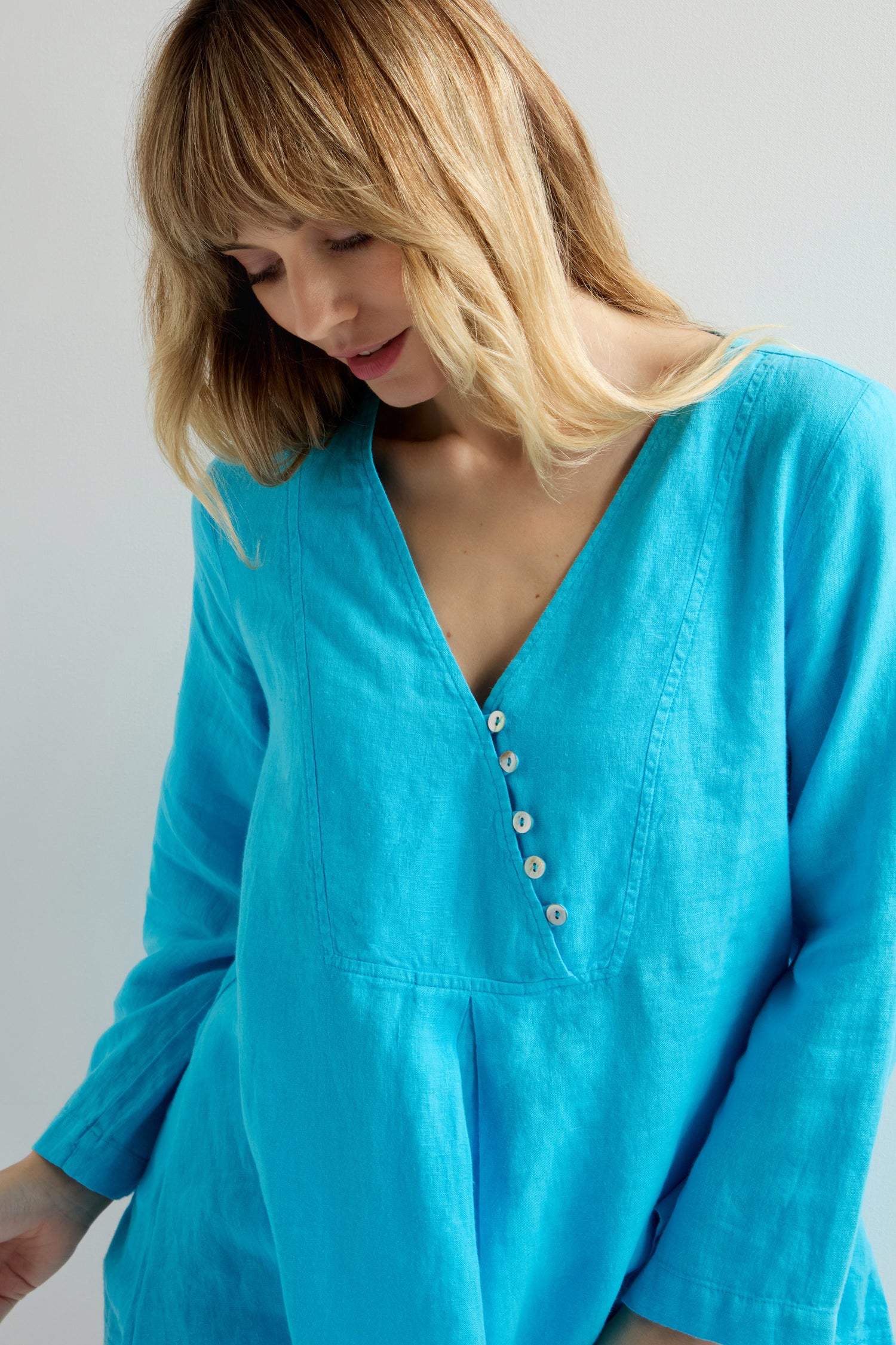Blonde individual in a turquoise Linen Button Placket Tunic, featuring a kaftan-inspired silhouette and buttons, looking down against a plain background.