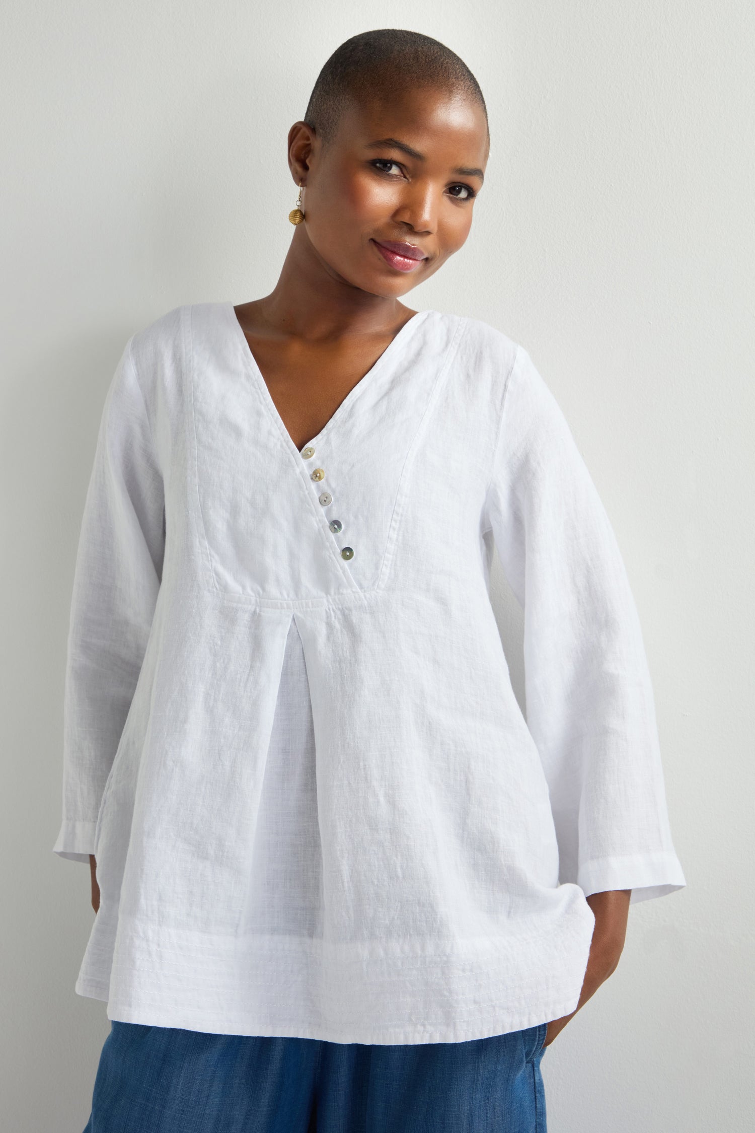 A person is standing against a white wall, wearing a relaxed silhouette V-neck top with buttons like the Linen Button Placket Tunic, paired with blue pants.