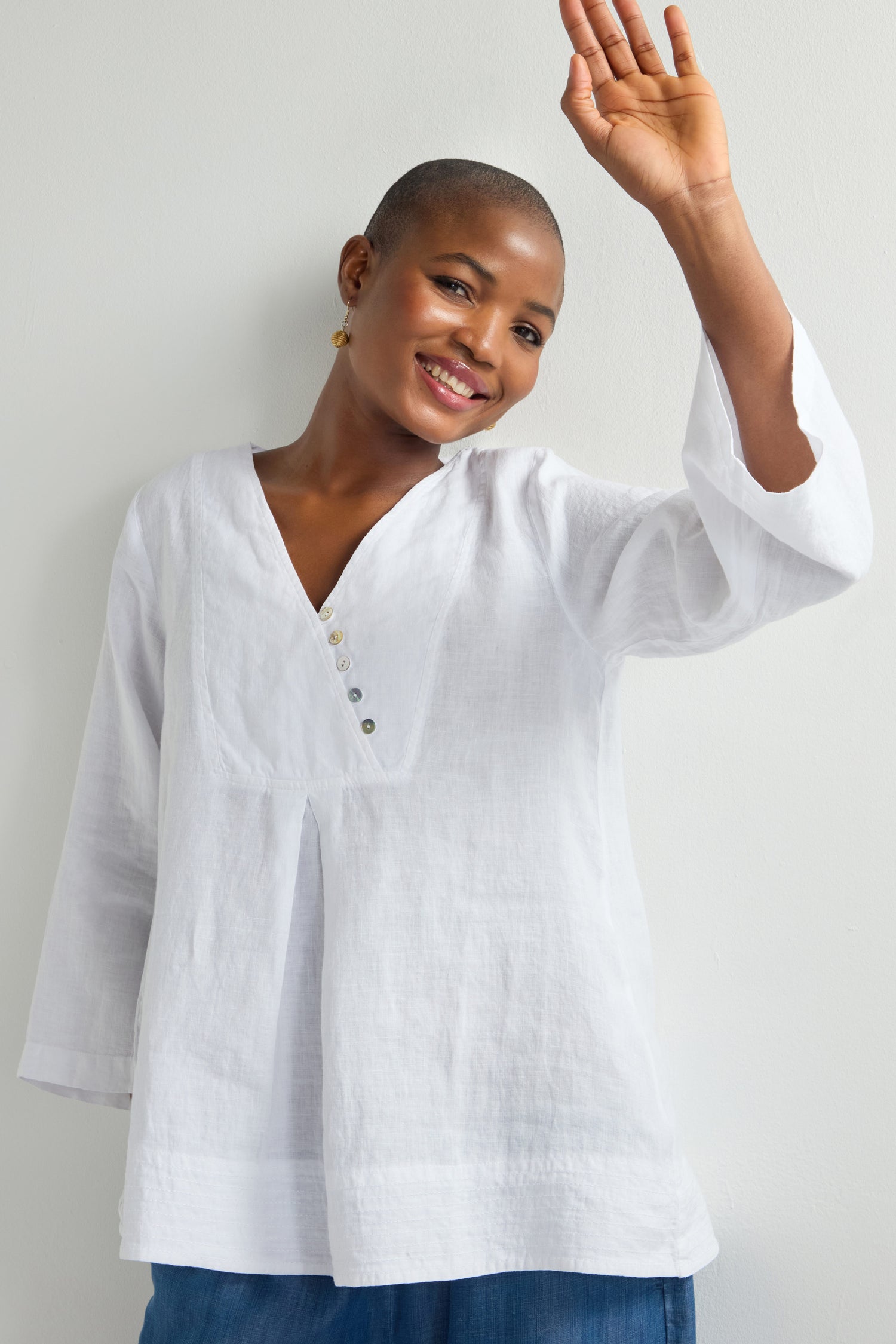 A person in a Linen Button Placket Tunic with a relaxed silhouette and blue pants smiles and waves against a plain background.