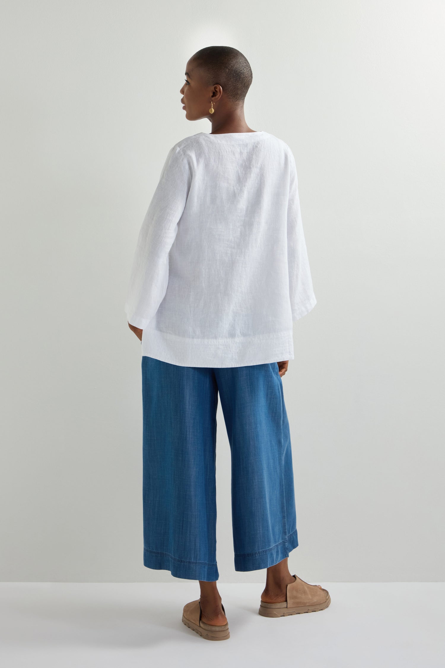 Someone in a white Linen Button Placket Tunic with a relaxed silhouette, blue wide-leg pants, and brown shoes stands on a light background, facing away.