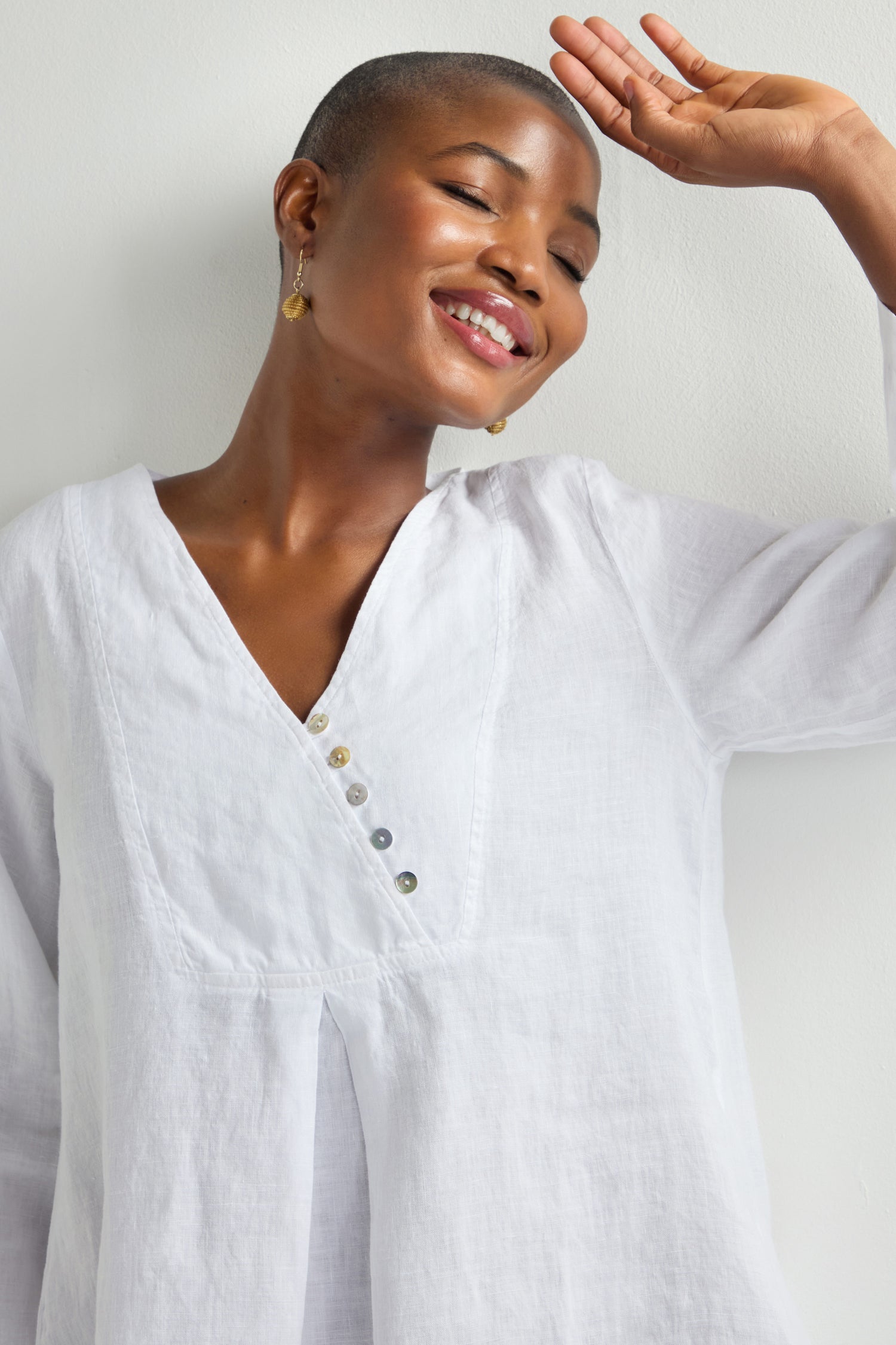 A person smiling, wearing a Linen Button Placket Tunic with a relaxed silhouette and gold earrings, their right arm raised against a white background.