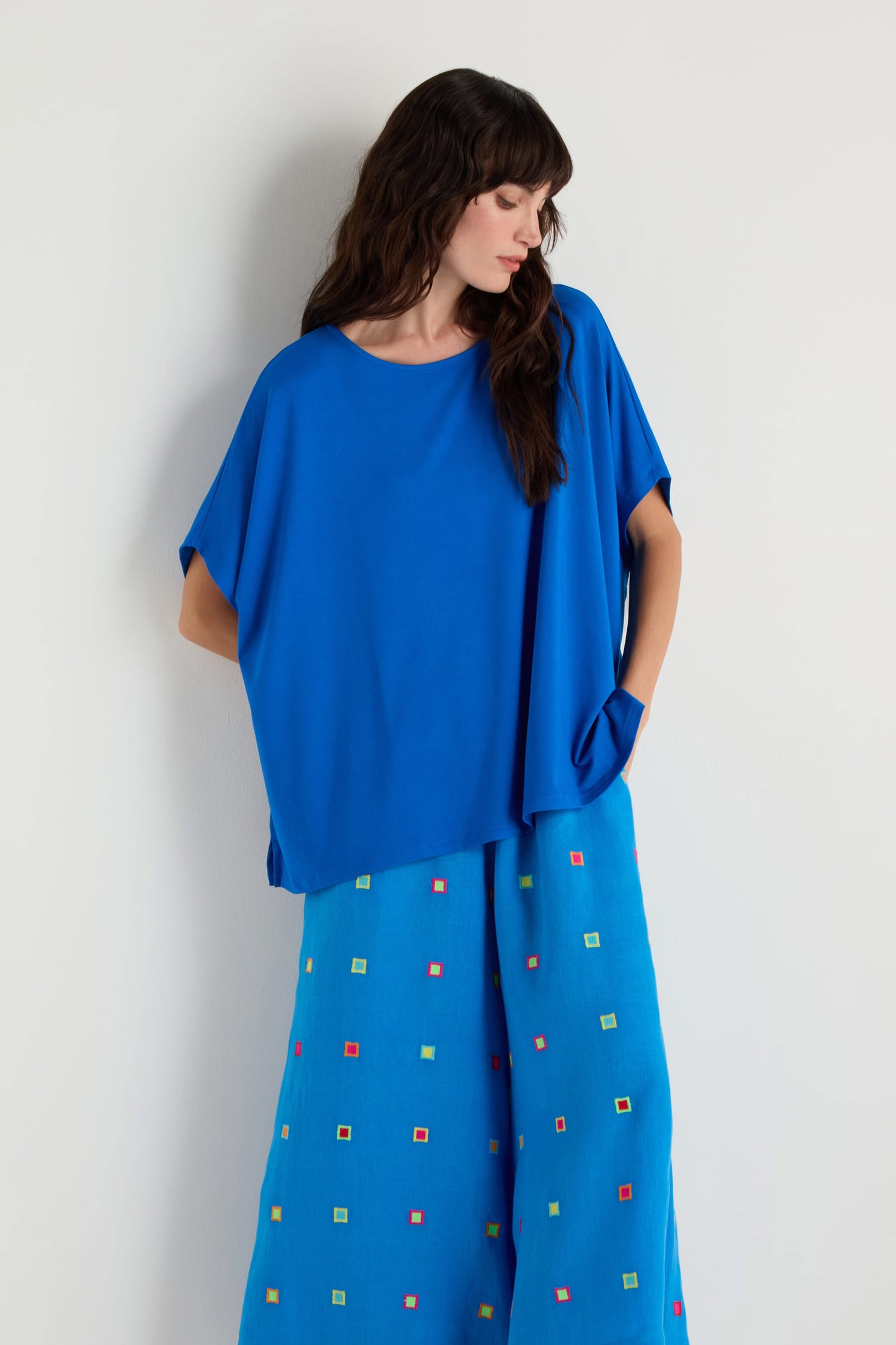 A woman wears the Viscose Jersey Boxy Top with wide-leg blue pants featuring small multicolored square patterns, standing against a plain off-white background.
