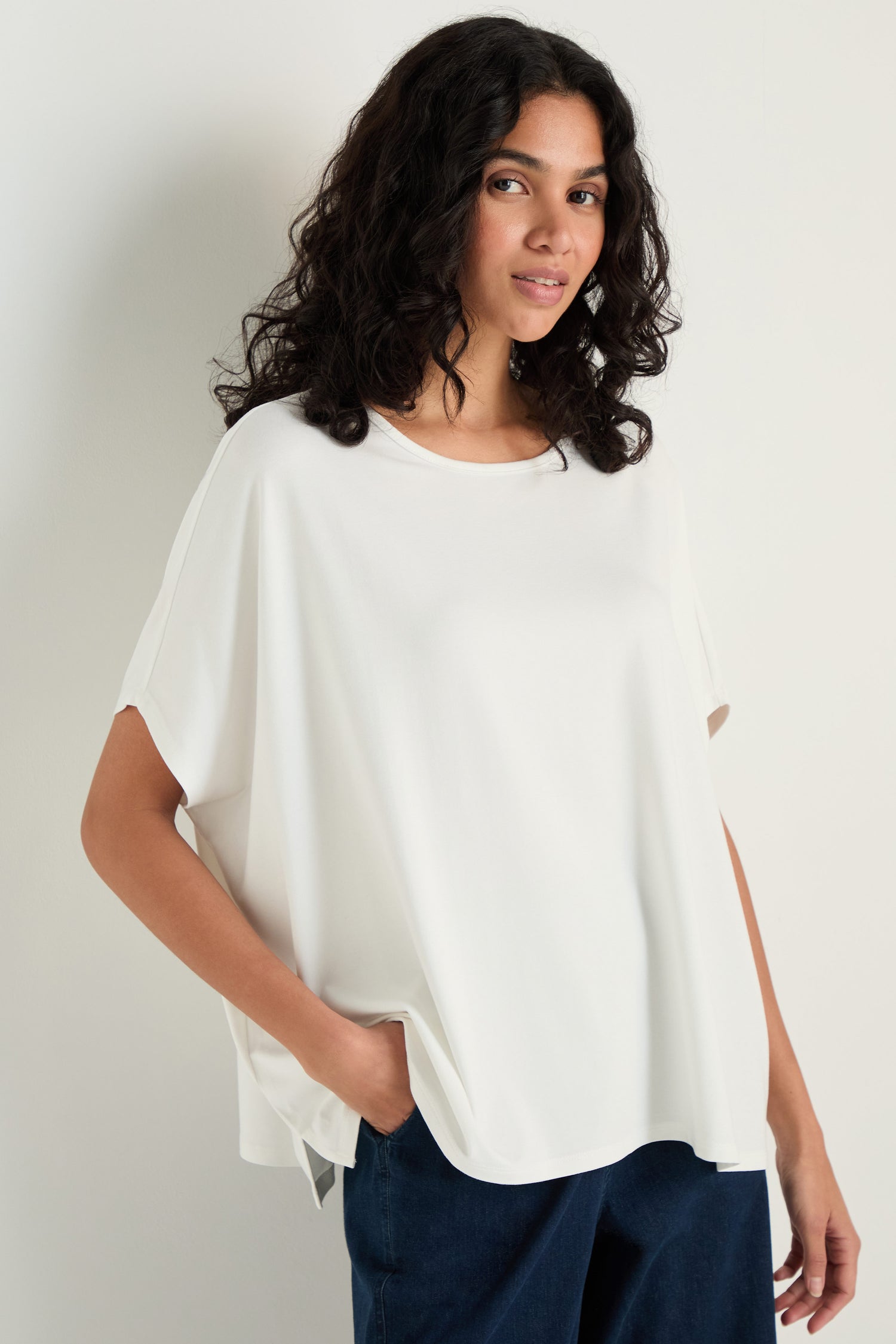 A woman with curly hair wears the Viscose Jersey Boxy Top and dark pants, standing against a plain light background with one hand in her pocket.