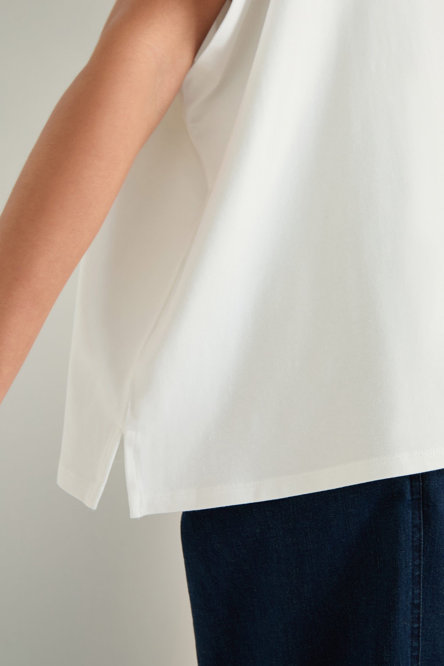 A person is shown from shoulder to thigh, wearing a white Viscose Jersey Boxy Top and dark blue pants, with one arm extended. The boxy silhouette of the top lends a modern edge to the minimalist ensemble.