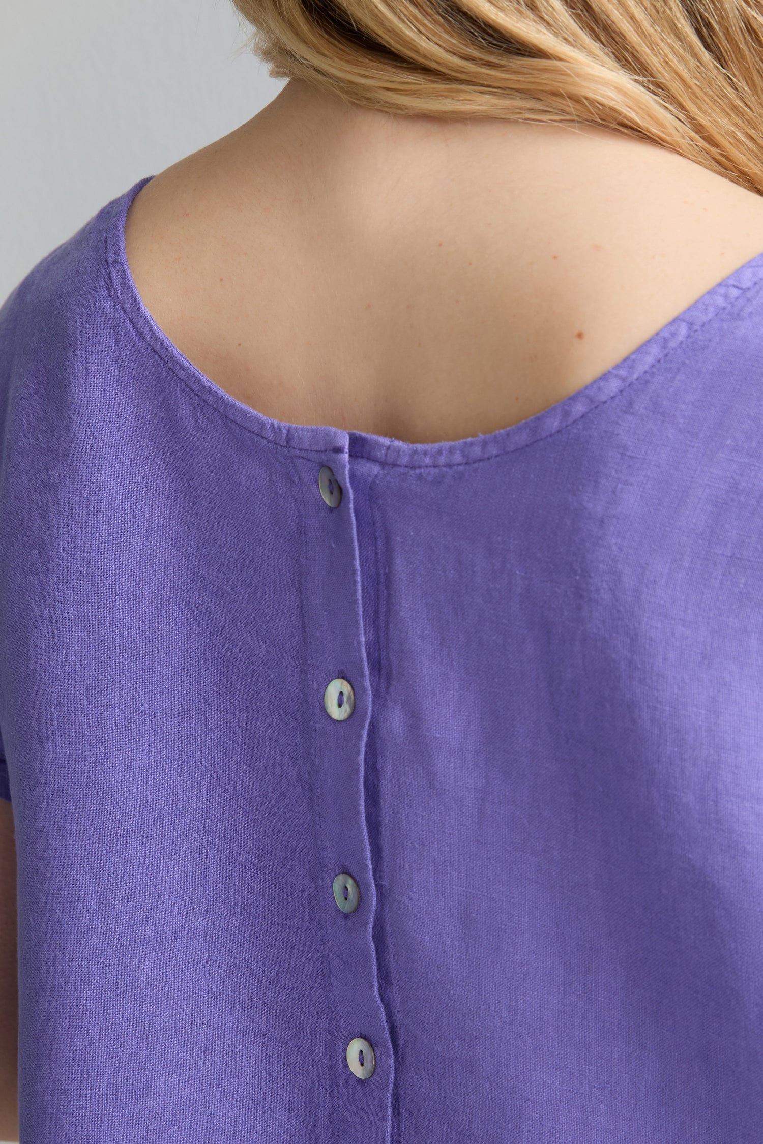 Close-up of a person wearing the Linen Button Back Top in purple, featuring an airy linen fabric, wide neckline, short sleeves, and buttons running down the back.