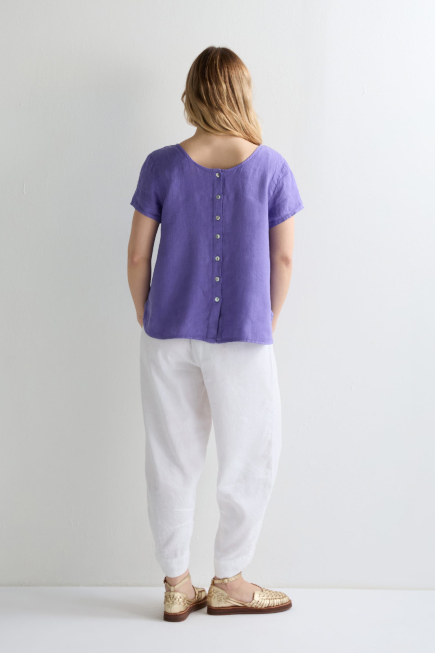 A woman faces a white wall, wearing the Linen Button Back Top in lavender with a subtle V neckline, paired with loose white pants and gold flat shoes.