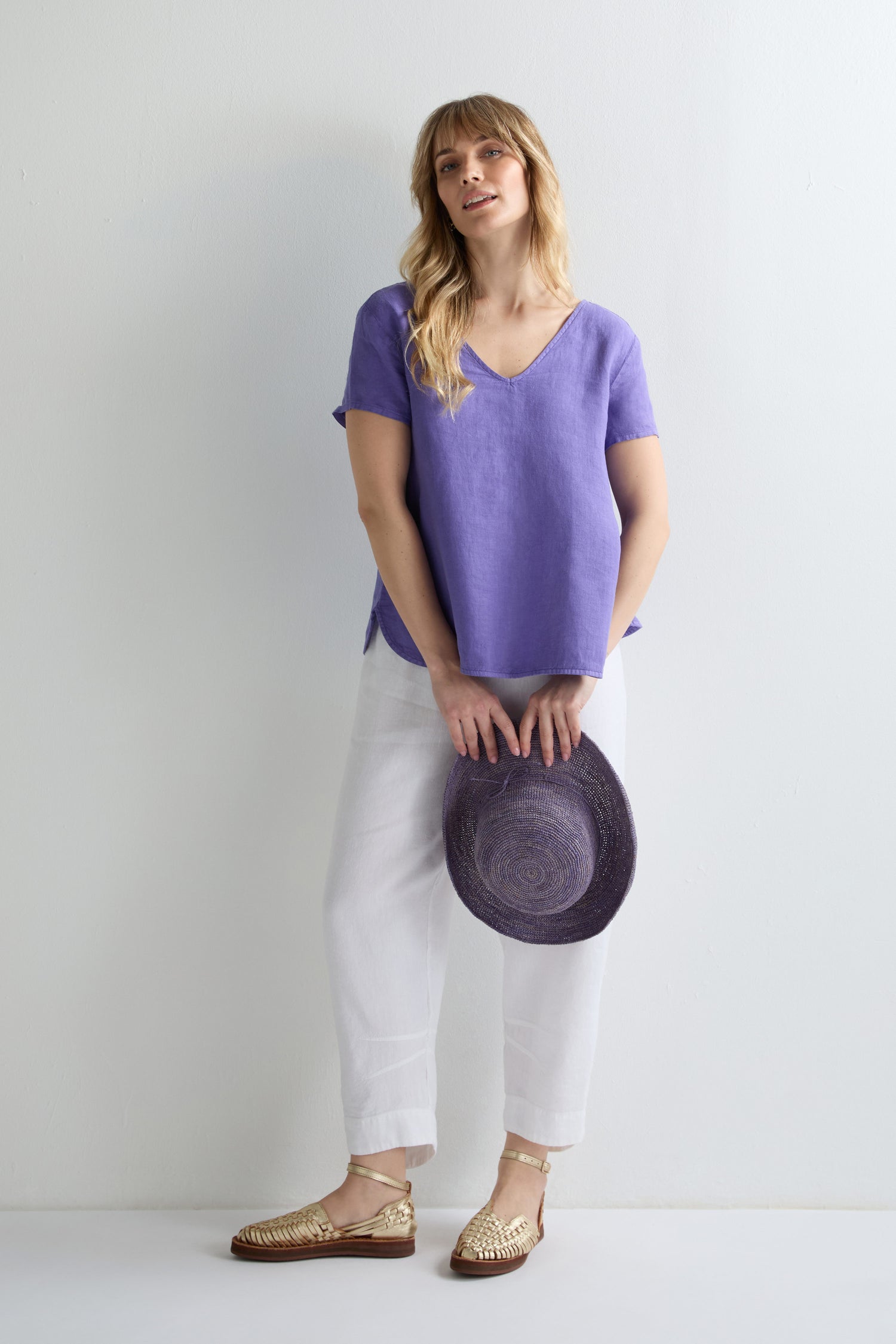 A woman stands against a white wall wearing the Linen Button Back Top in purple, paired with white pants, gold sandals, and holding a matching purple hat.