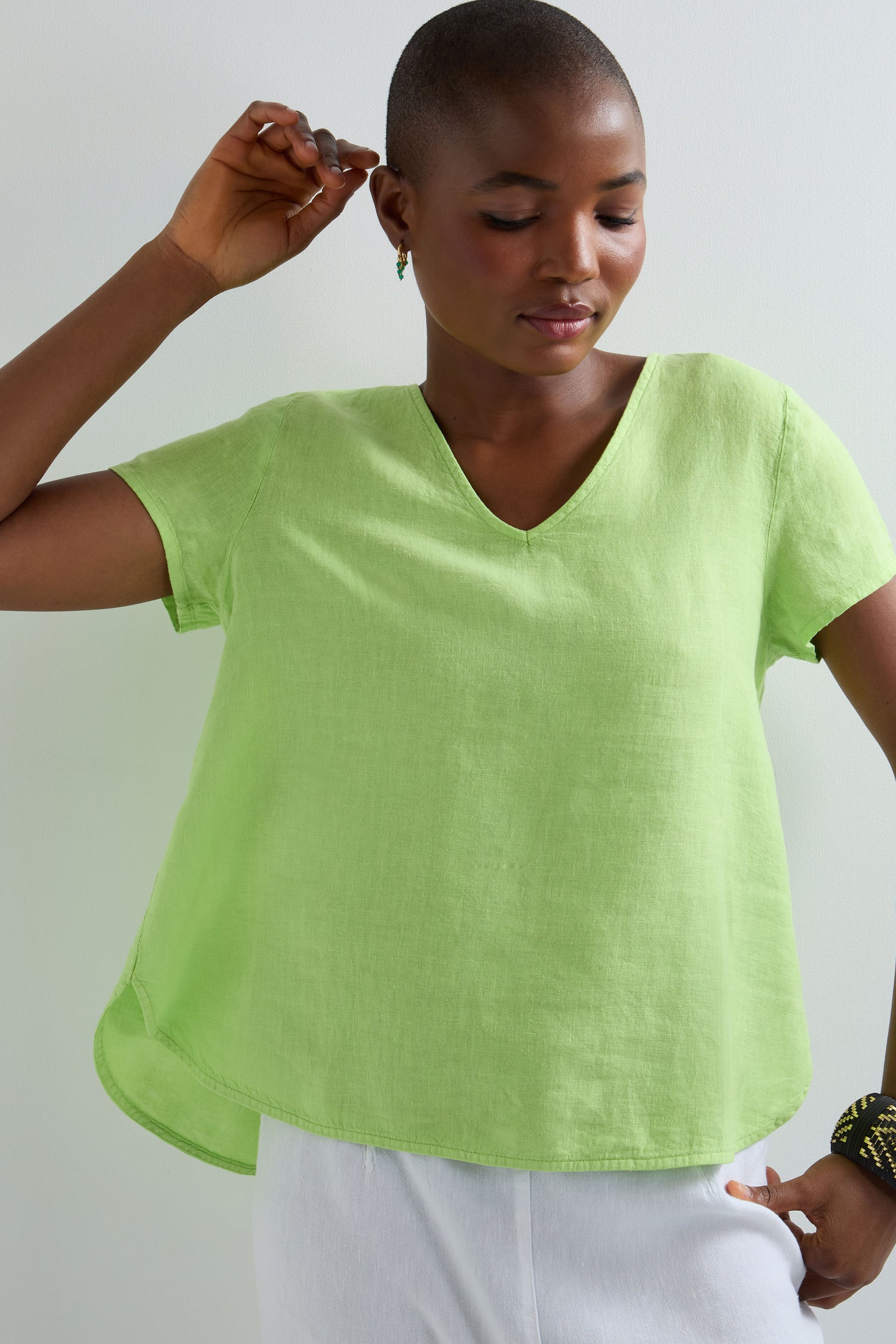 A woman in a light green Garment Dyed Linen Button Back Top with short sleeves and a V neckline pairs it with white pants, standing against a plain light background and slightly raising one arm.