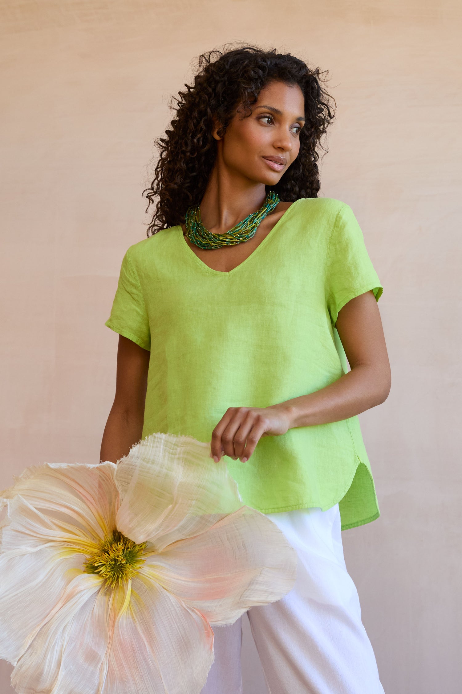 A woman in a lime green Garment Dyed Linen Button Back Top and white pants holds a large artificial flower, standing against a neutral background. She accessorizes with a layered green necklace.
