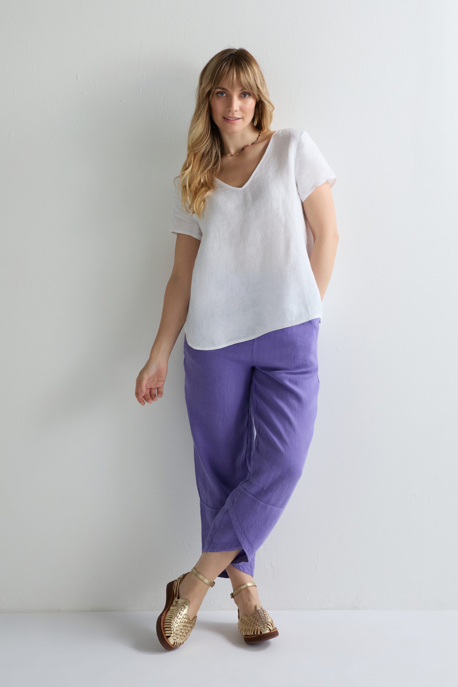 A woman stands against a white wall, wearing a Linen Button Back Top with a V neckline, loose purple pants, and gold flat sandals.
