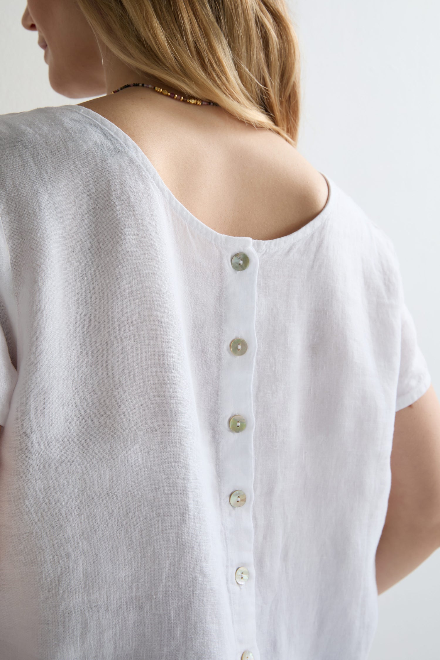 A person models the Linen Button Back Top, featuring a breezy linen fabric, button-back detail, and V neckline, with a glimpse of their shoulder and neck visible.