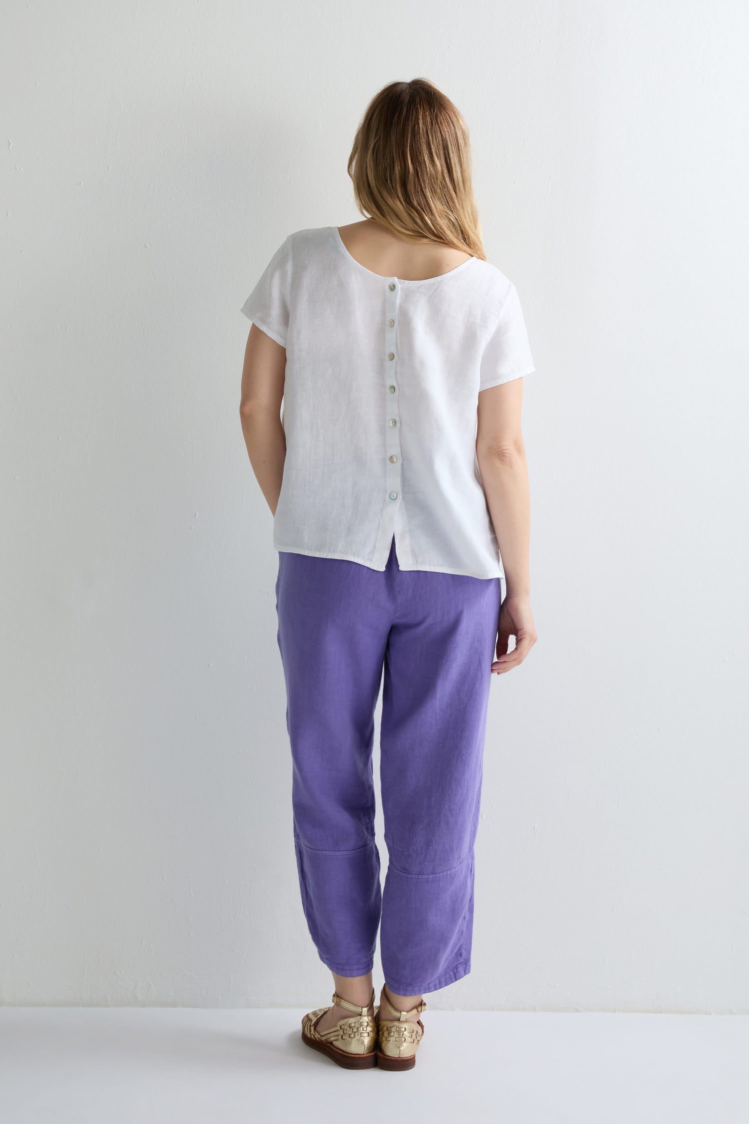 A woman stands facing a white wall, wearing the Linen Button Back Top with a V neckline, paired with purple pants and beige sandals.