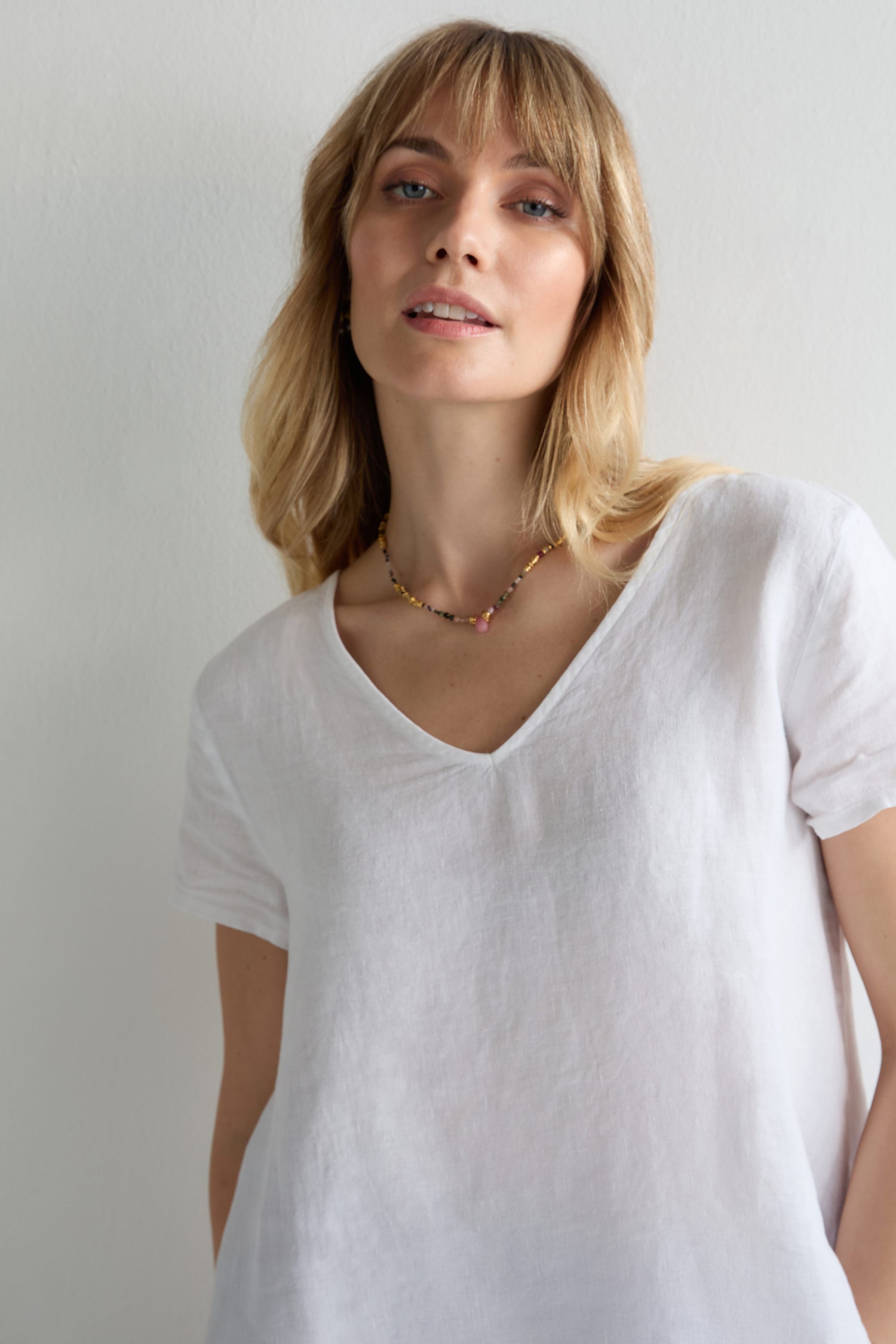 A woman with blonde hair wears the Linen Button Back Top and a beaded necklace as she stands in front of a plain, light-colored background.