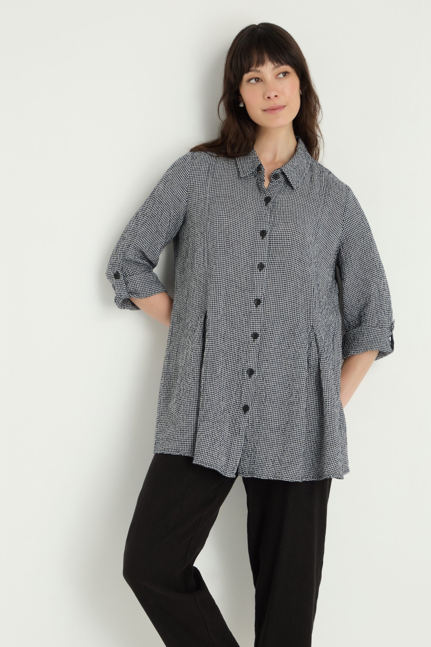A woman stands against a plain white background wearing the Micro Gingham Shirt—loose-fitting with long sleeves in black and white—paired with black pants.