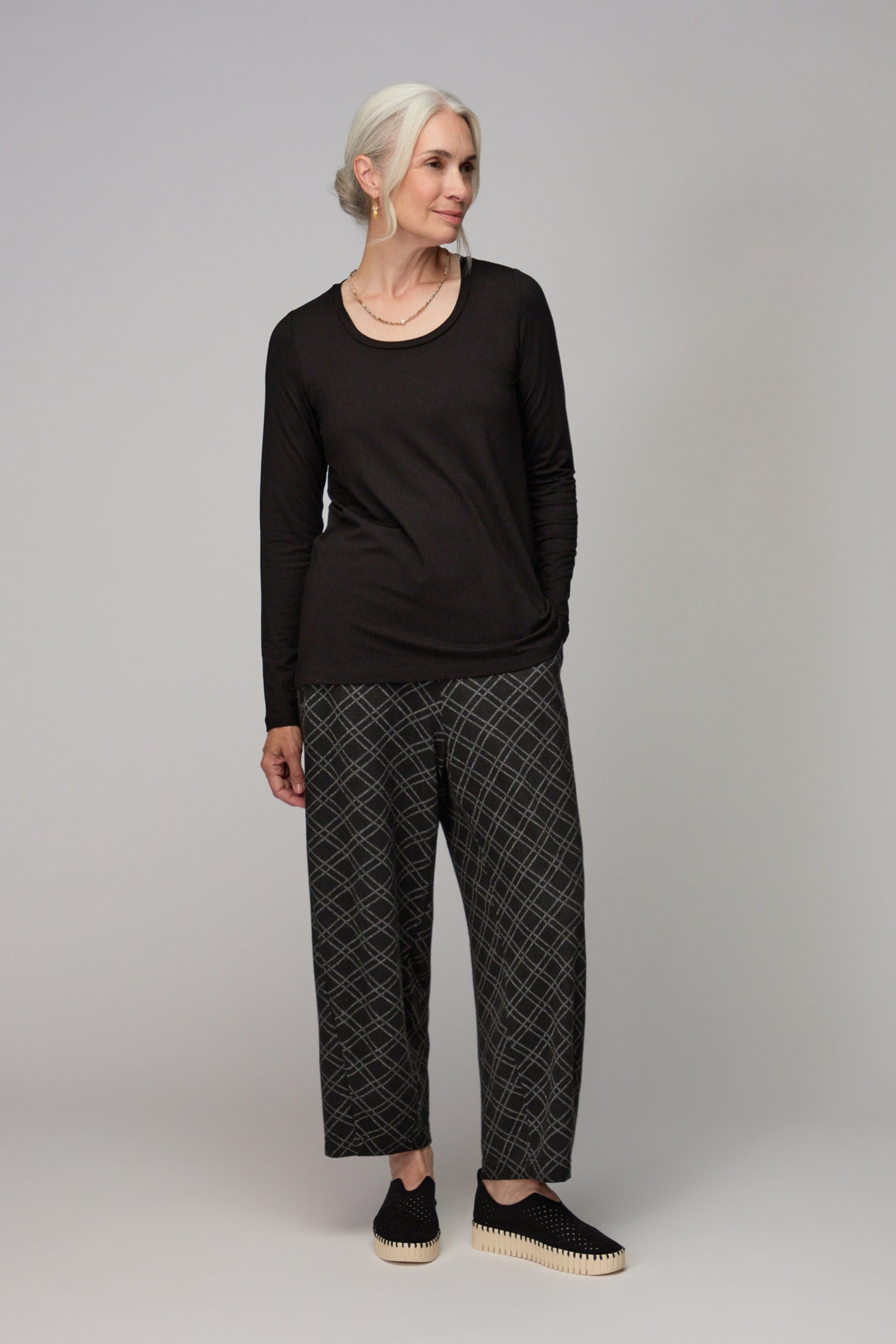 A gray-haired woman wears the Viscose Jersey Scoop Neck Top in black, paired with patterned wide-leg pants and black slip-on shoes, standing against a plain gray background.