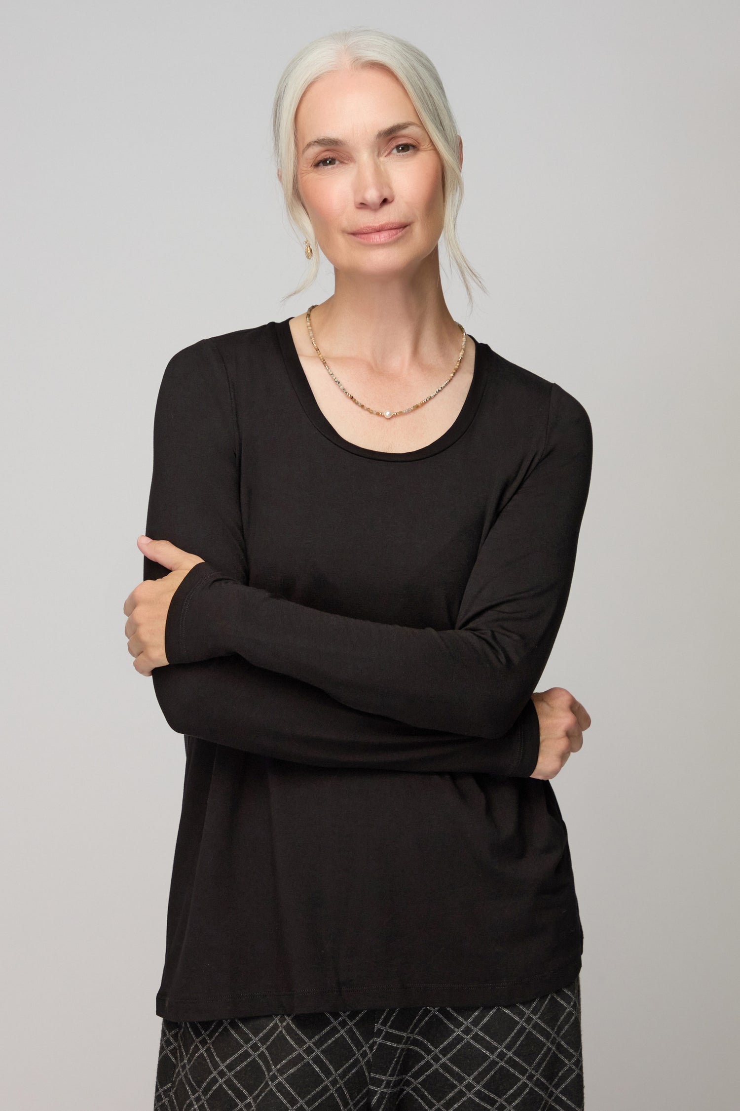 A woman with gray hair wears the Viscose Jersey Scoop Neck Top in black and checkered pants, standing with arms crossed and looking at the camera against a plain background.
