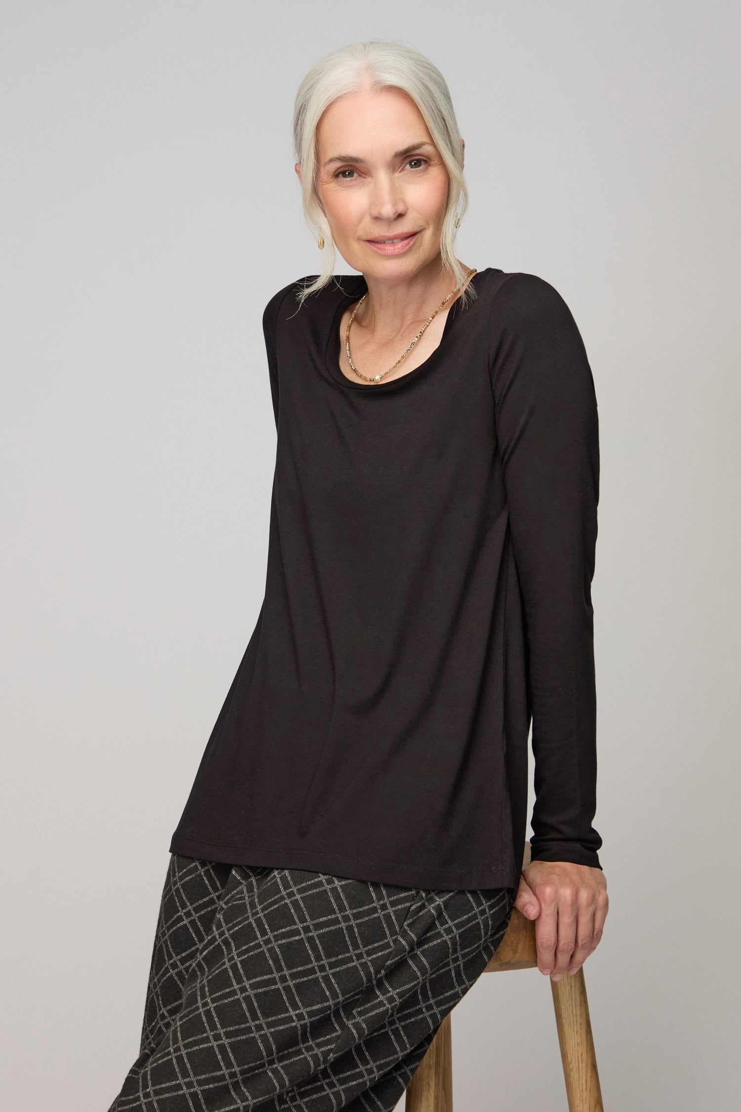 An older woman with gray hair sits on a wooden stool against a plain light background, looking at the camera while wearing a slim fit, long-sleeve black Viscose Jersey Scoop Neck Top and a plaid skirt.