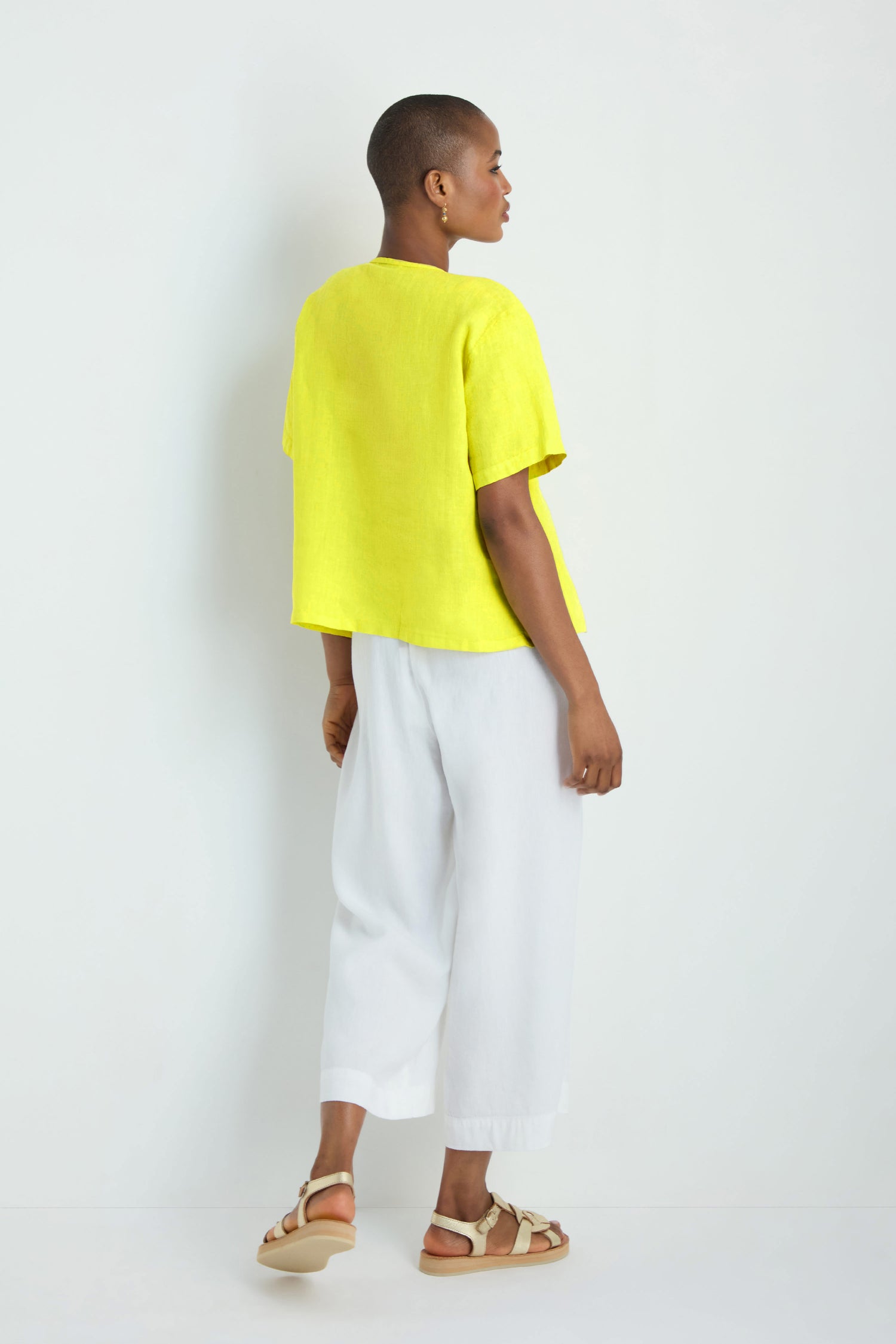 A person stands against a plain white background, wearing the Linen Pleat Short Sleeve Lemon Shirt, paired with wide-leg white pants and beige sandals, facing slightly away from the camera.