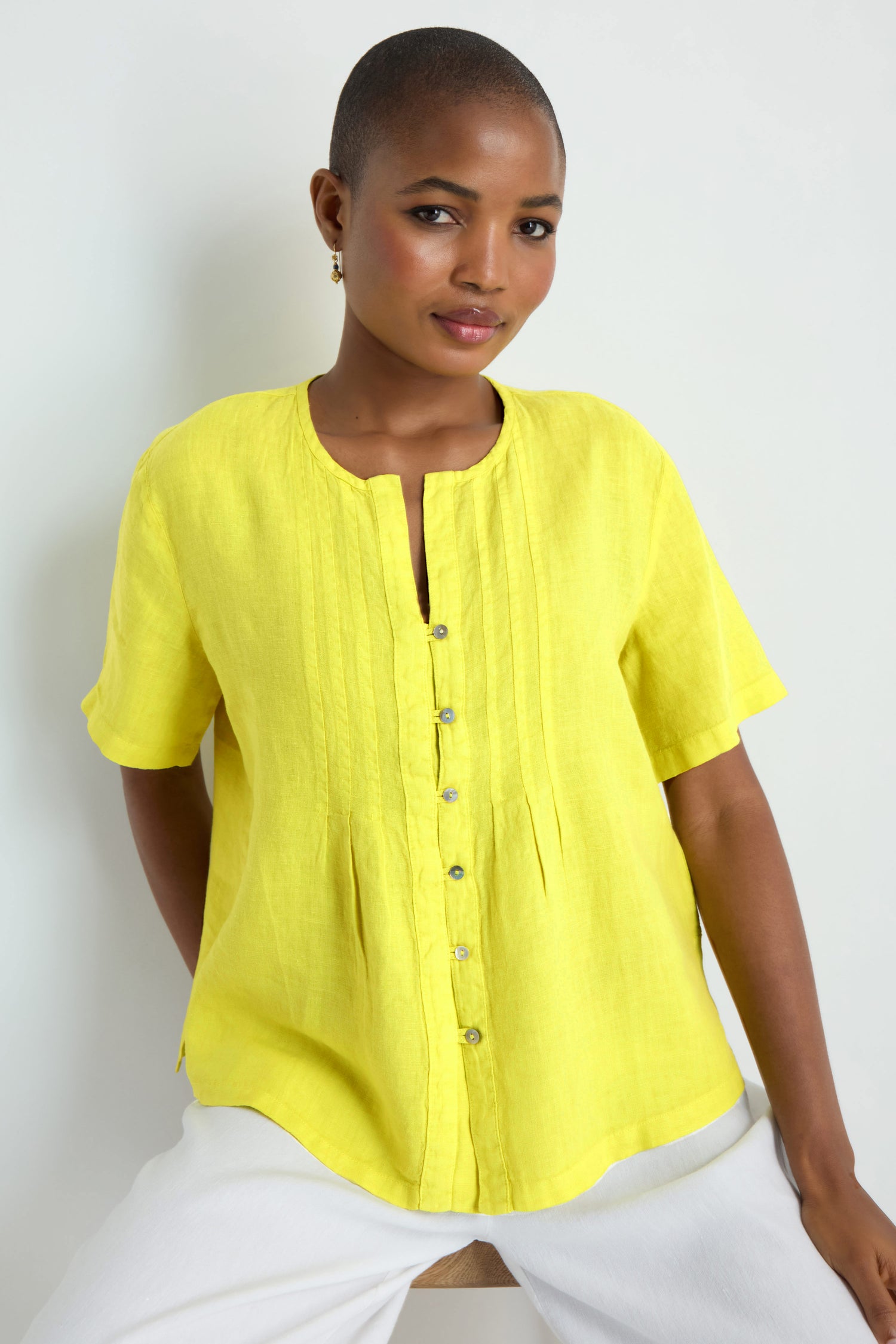 A person poses against a plain white background wearing the Linen Pleat Short Sleeve Lemon Shirt—lightweight, short-sleeved, button-up in yellow—paired with white pants.
