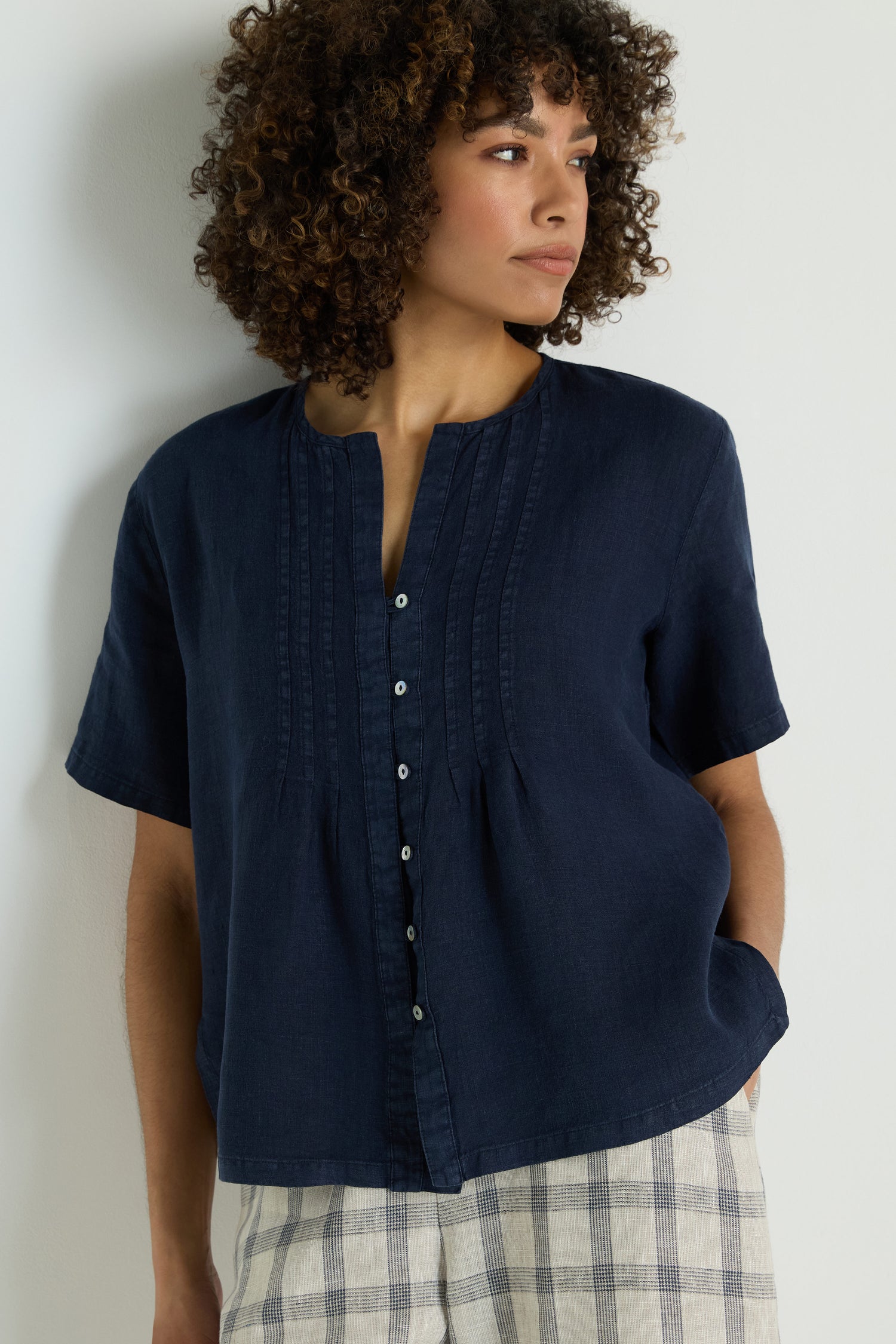A woman wearing the Linen Pleat Short Sleeve Shirt in navy blue and light gray pants stands against a plain white background, looking down with one hand in her pocket.