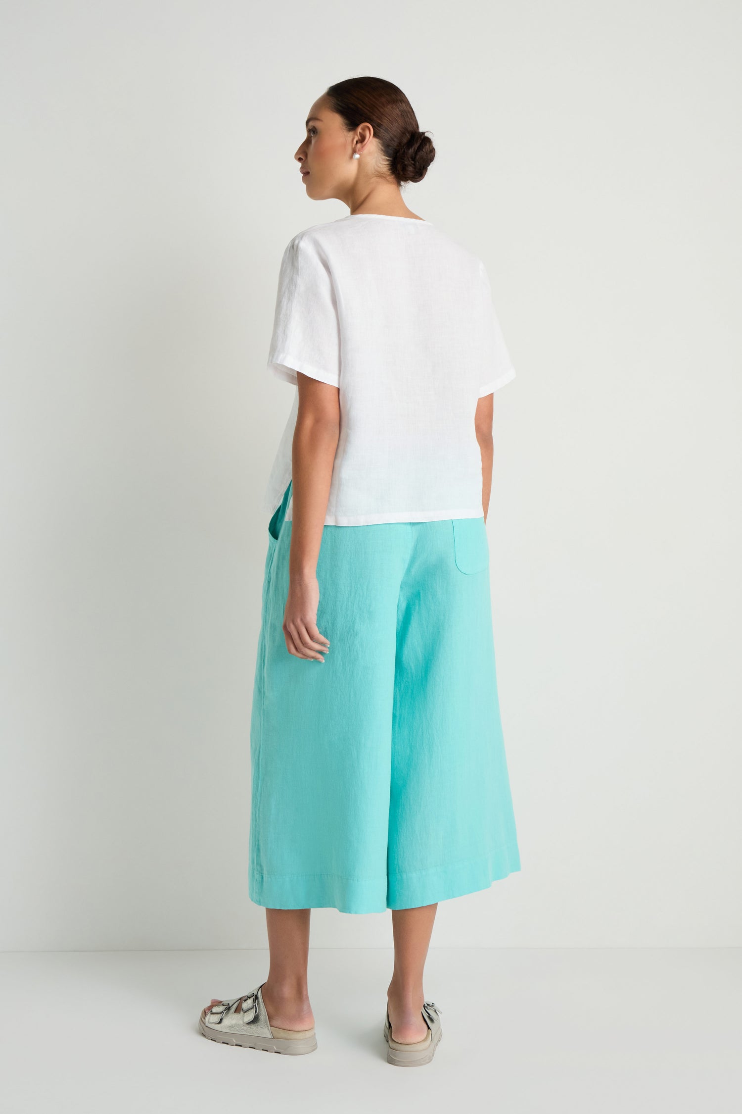 A woman faces away, wearing the Linen Pleat Short Sleeve White Shirt, aqua culotte pants, and beige sandals against a plain white background.