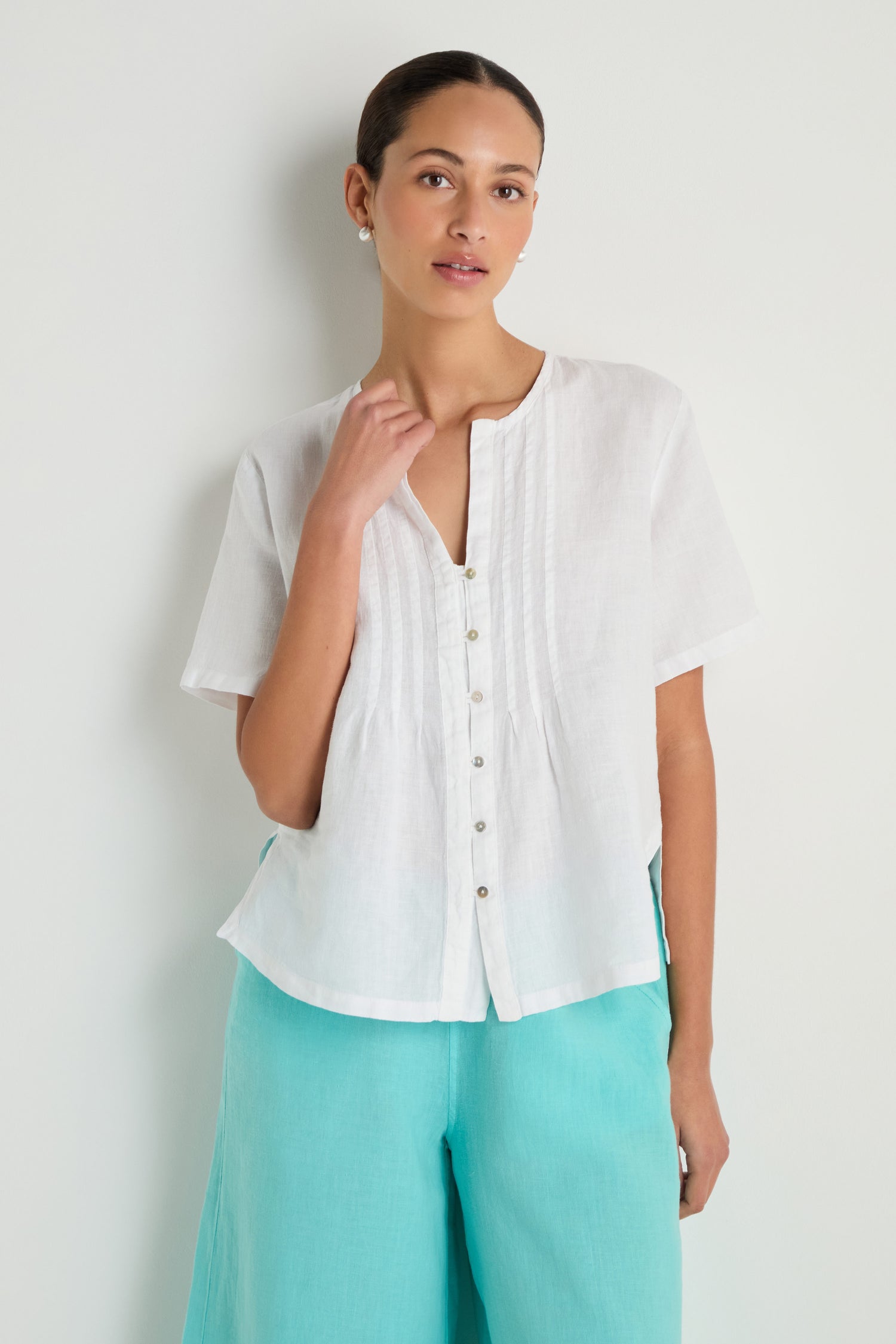 A woman wears the Linen Pleat Short Sleeve White Shirt with light turquoise pants, standing against a plain light background.