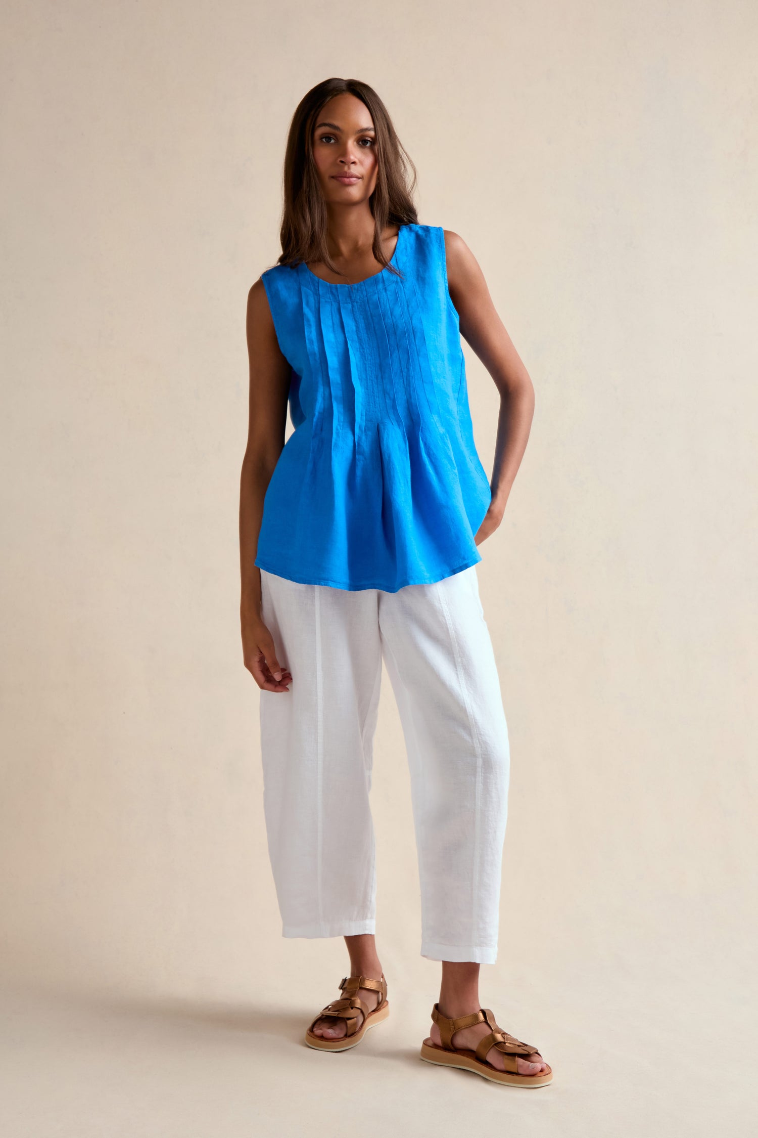 A woman stands against a plain background wearing the Linen Pleat Front Azure Vest, paired with white loose-fitting pants and brown sandals.