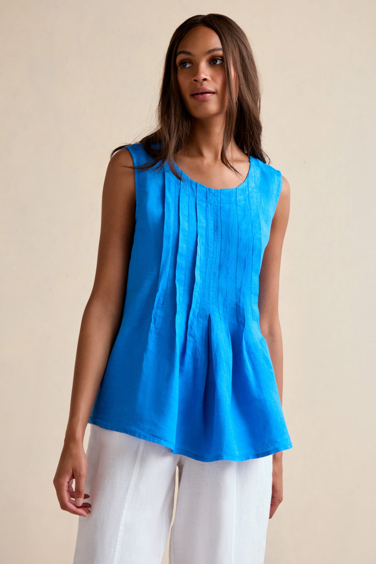 A woman wears the Linen Pleat Front Azure Vest, a sleeveless bright blue linen top with pleats, paired with white pants against a light beige background.