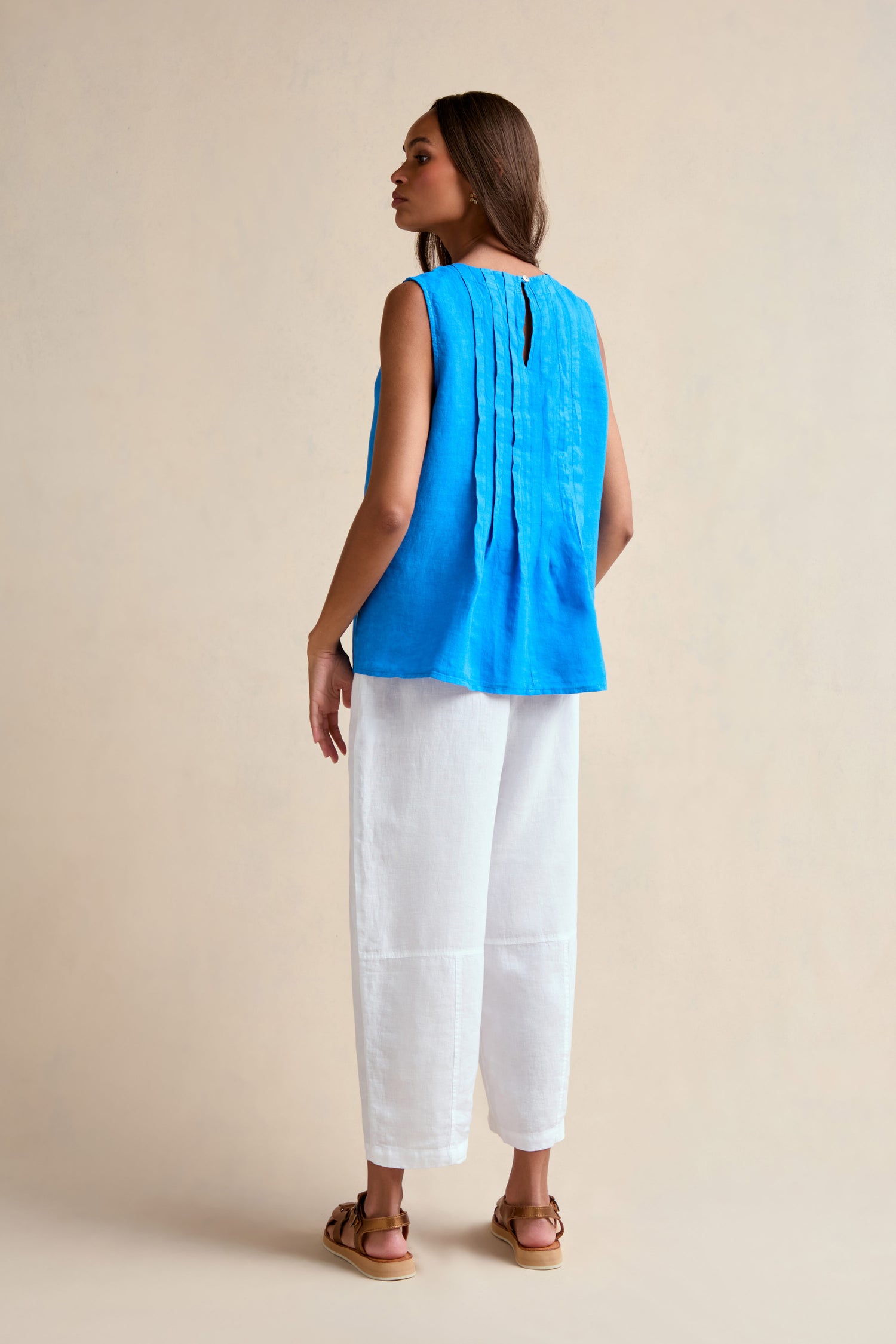 A woman stands with her back to the camera, wearing the Linen Pleat Front Azure Vest with vertical pleats, loose white pants, and brown sandals against a neutral background.