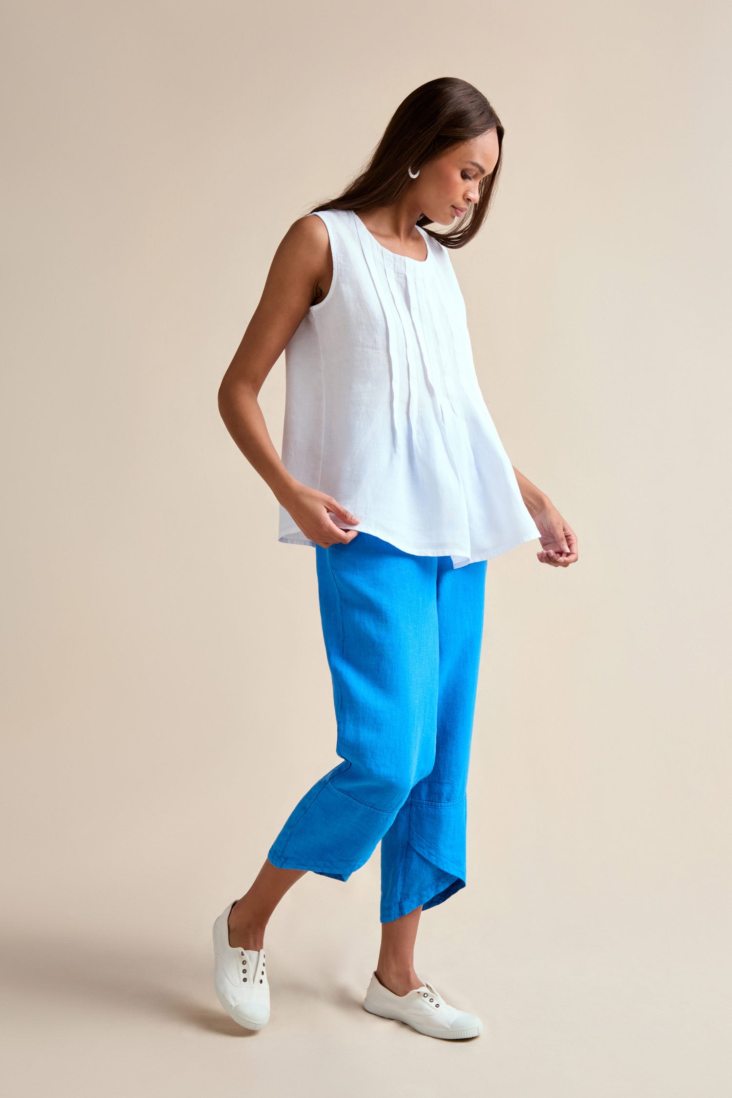 A woman stands in a neutral space wearing the Linen Pleat Front White Vest, bright blue pleat front cropped pants, and white sneakers.