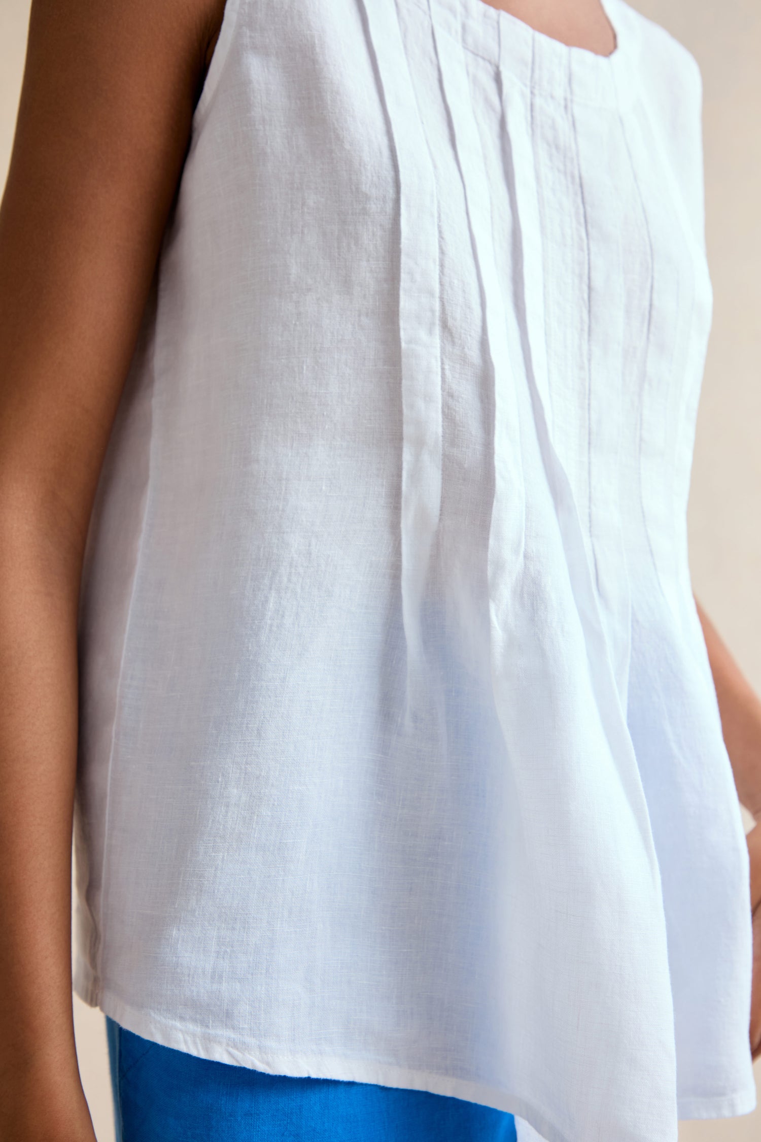 Someone is wearing the Linen Pleat Front White Vest, which is sleeveless and features pleat front details, styled with bright blue pants.