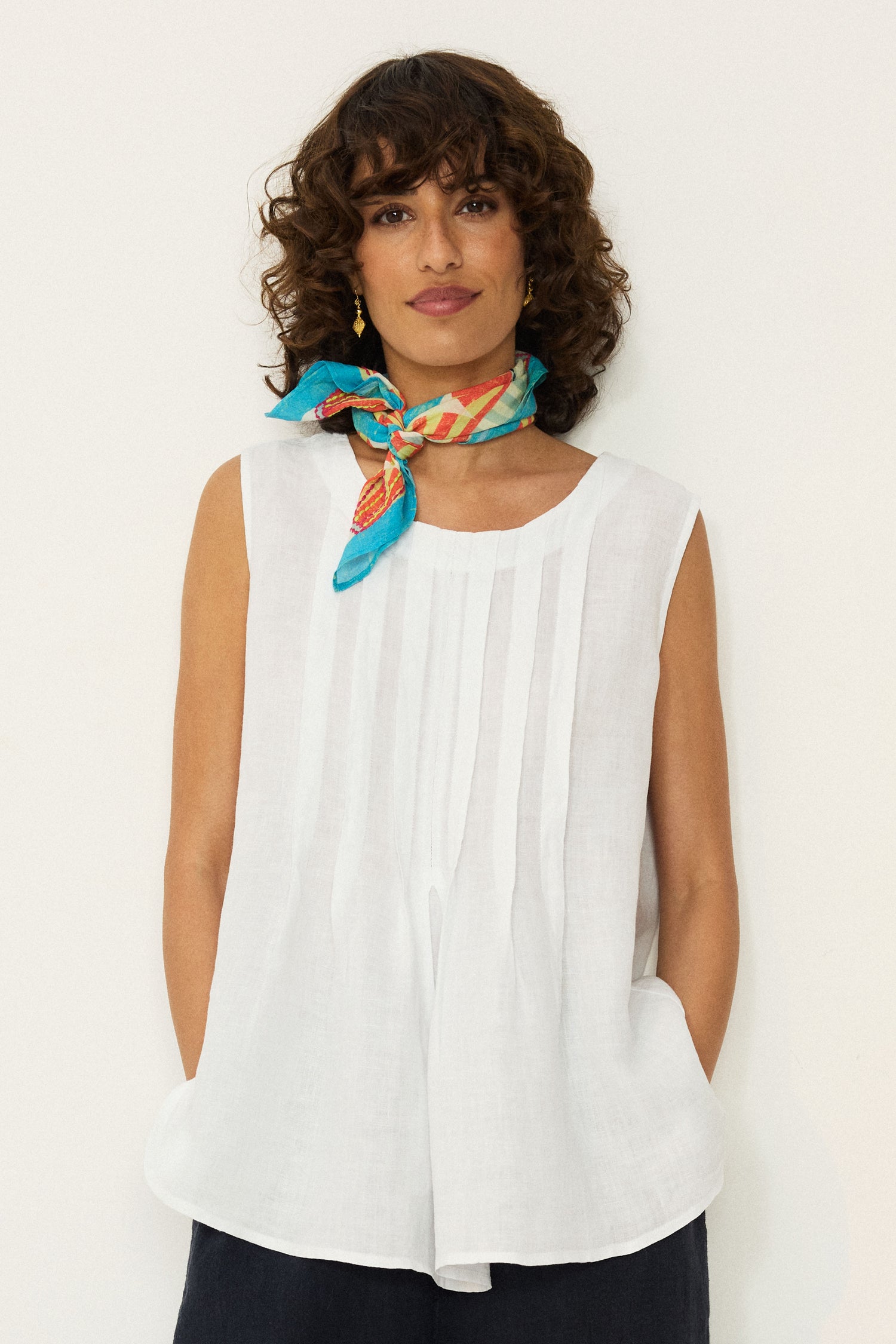 A woman with curly hair wears the Linen Pleat Front White Vest and a colorful scarf, standing against a plain white background with her hands in her pockets.