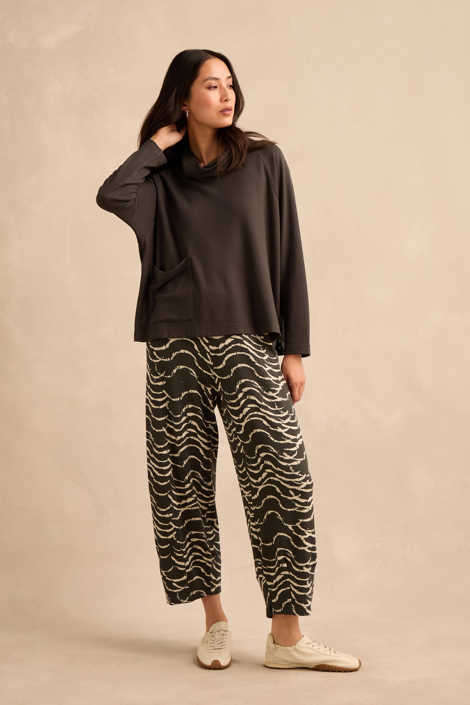 A woman stands against a beige background, wearing the Luxe Viscose Jersey Cowl Neck Top, patterned wide-leg pants, and white sneakers, with one hand touching her neck.