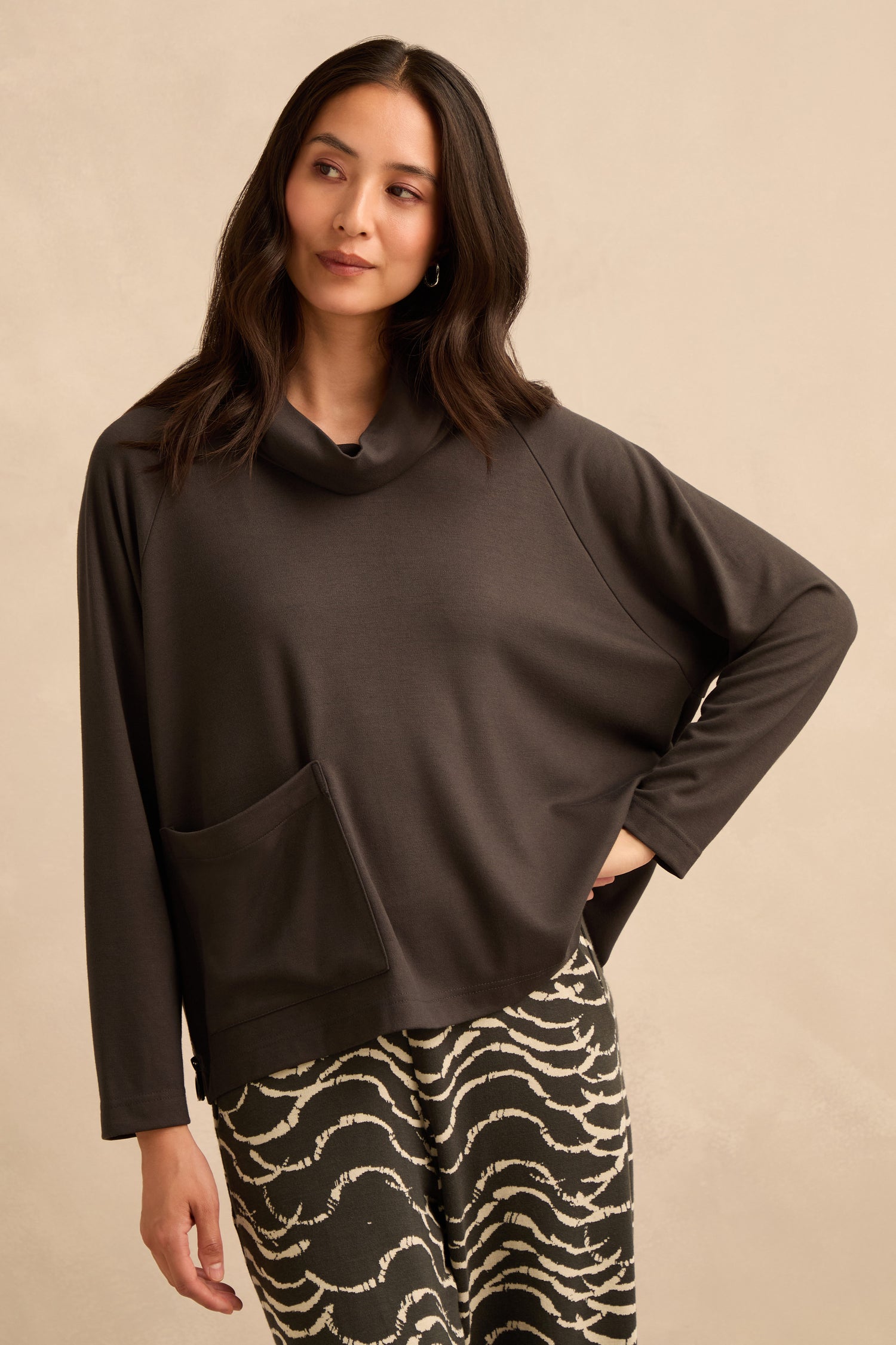A woman wearing a Luxe Viscose Jersey Cowl Neck Top with long sleeves and a front pocket, paired with patterned black and white pants, stands against a beige background.